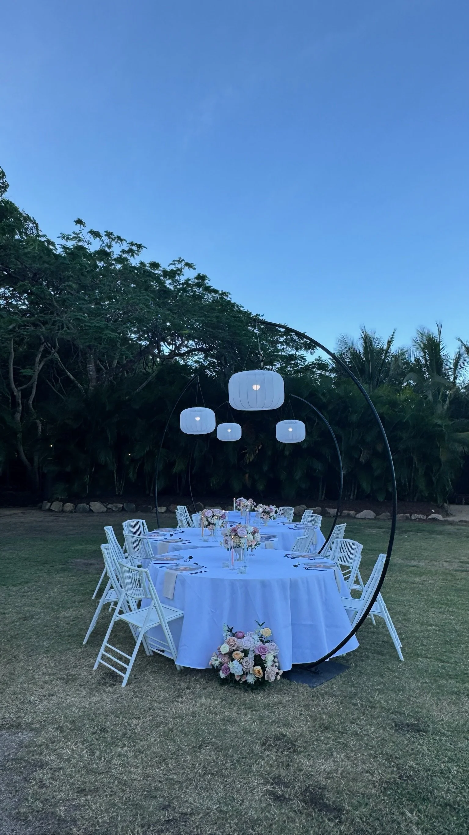 Outdoor dining setup with a large round table covered with a white tablecloth, decorated with flower arrangements, surrounded by white chairs, under hanging white lanterns, on a grassy area with trees in the background.