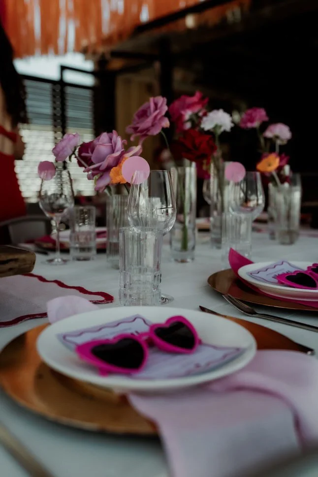 A table set for a celebration with pink and purple flowers in vases, pink and black heart-shaped sunglasses on plates, and glasses of water. The setting appears to be indoors with wooden and windowed background.