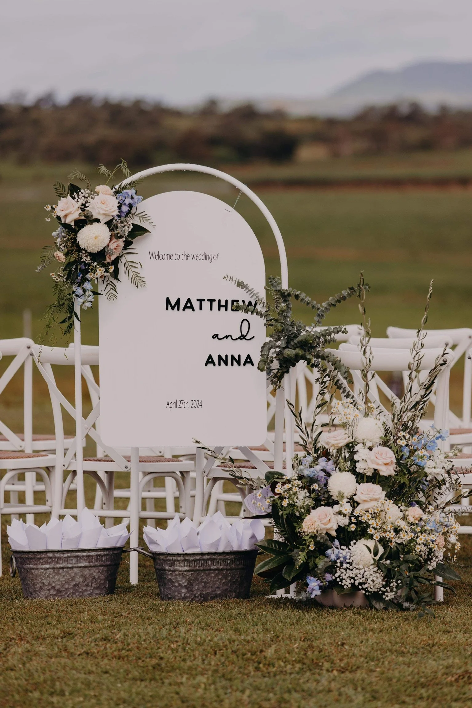 Wedding sign reading 'Welcome to the wedding of MATTHEW and ANNA' with the date April 27, 2024, decorated with floral arrangements and greenery, set on chairs in an outdoor setting with green grass and a blurred landscape in the background.