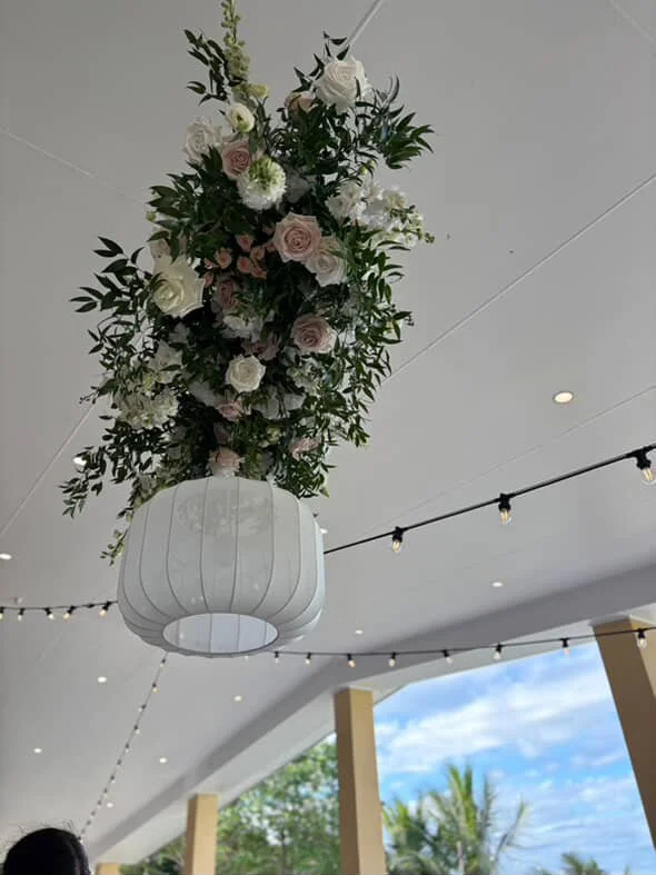 Hanging floral arrangement with pink and white roses and greenery in a white lantern-style vase, ceiling with string lights and outdoor view with blue sky and trees.