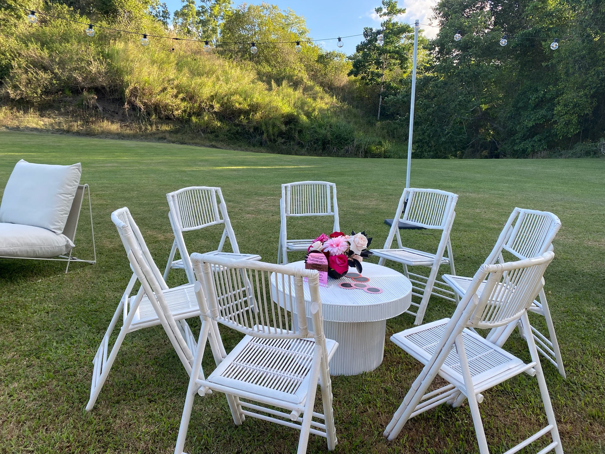 Outdoor patio setup with a round white table surrounded by eight white chairs, pink and white flowers in a vase on the table, and a large white cushioned lounge chair on the grass. String lights hang overhead in a green backyard with trees and a hill