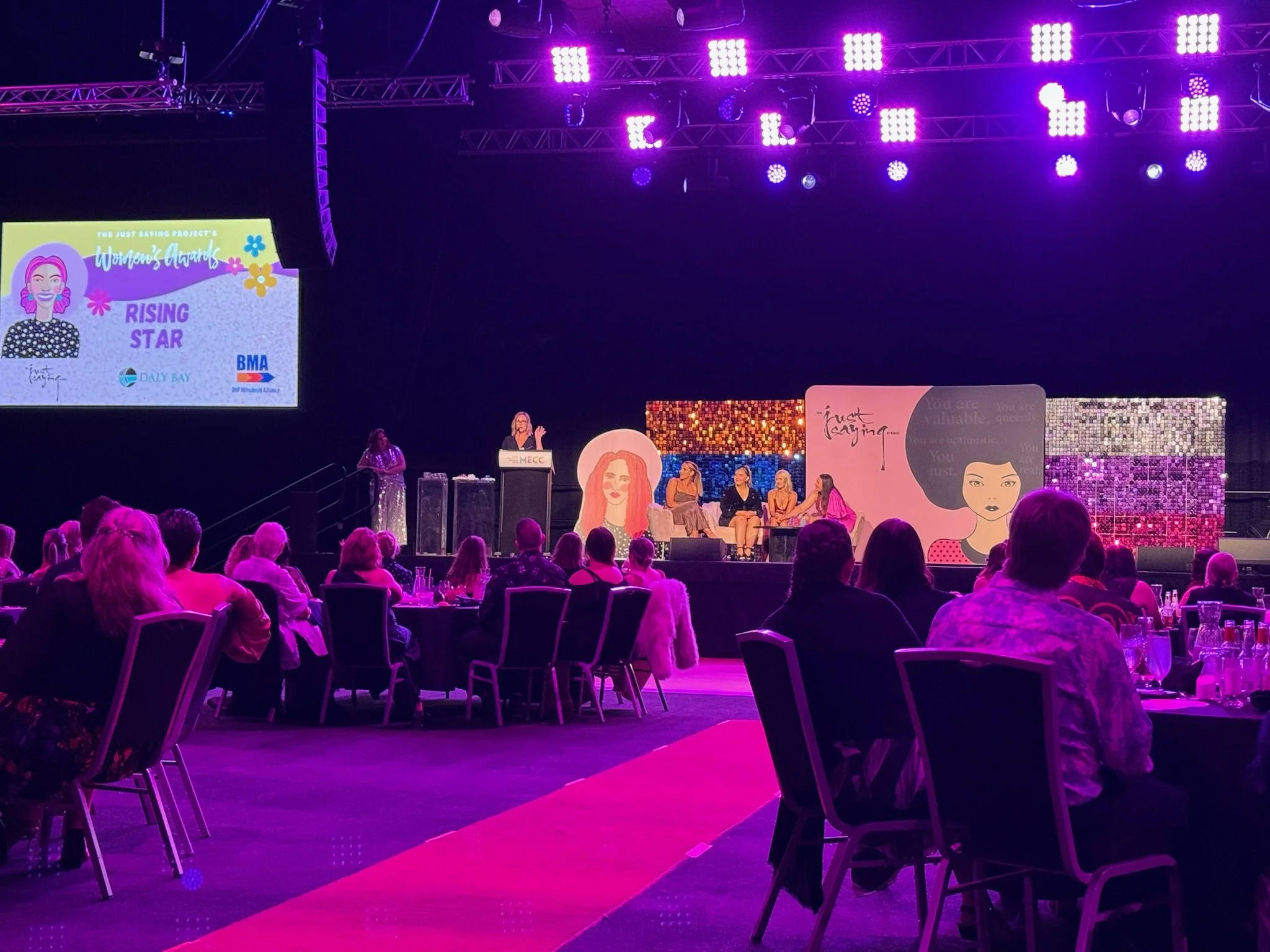 An indoor award ceremony with a stage, purple lighting, and an audience seated at round tables. On the stage, a woman is speaking at a podium, with four women seated behind her. A large screen displays the event title and logos. The background featur