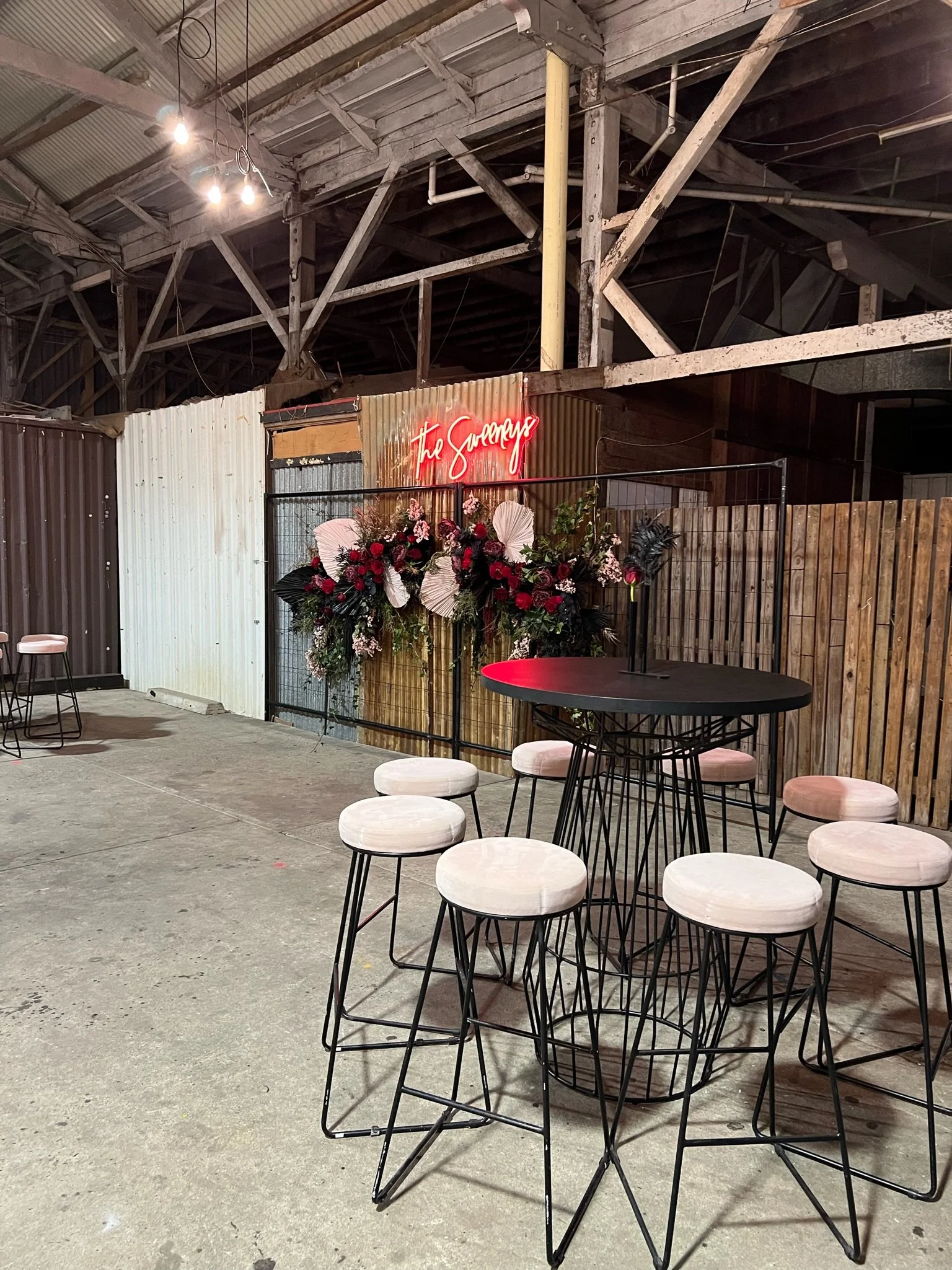 Industrial-style event space with a round black table surrounded by eight cream-colored stools with thin black metal legs. In the background, there is a floral arrangement with red, white, and dark green flowers and a neon sign that reads "The Savage