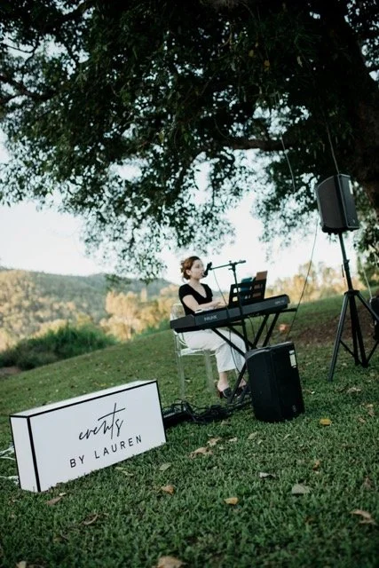 A woman playing a keyboard and singing outdoors under a large tree, with a speaker and a sign that reads 'Wenty by Lauren', in a scenic park setting.