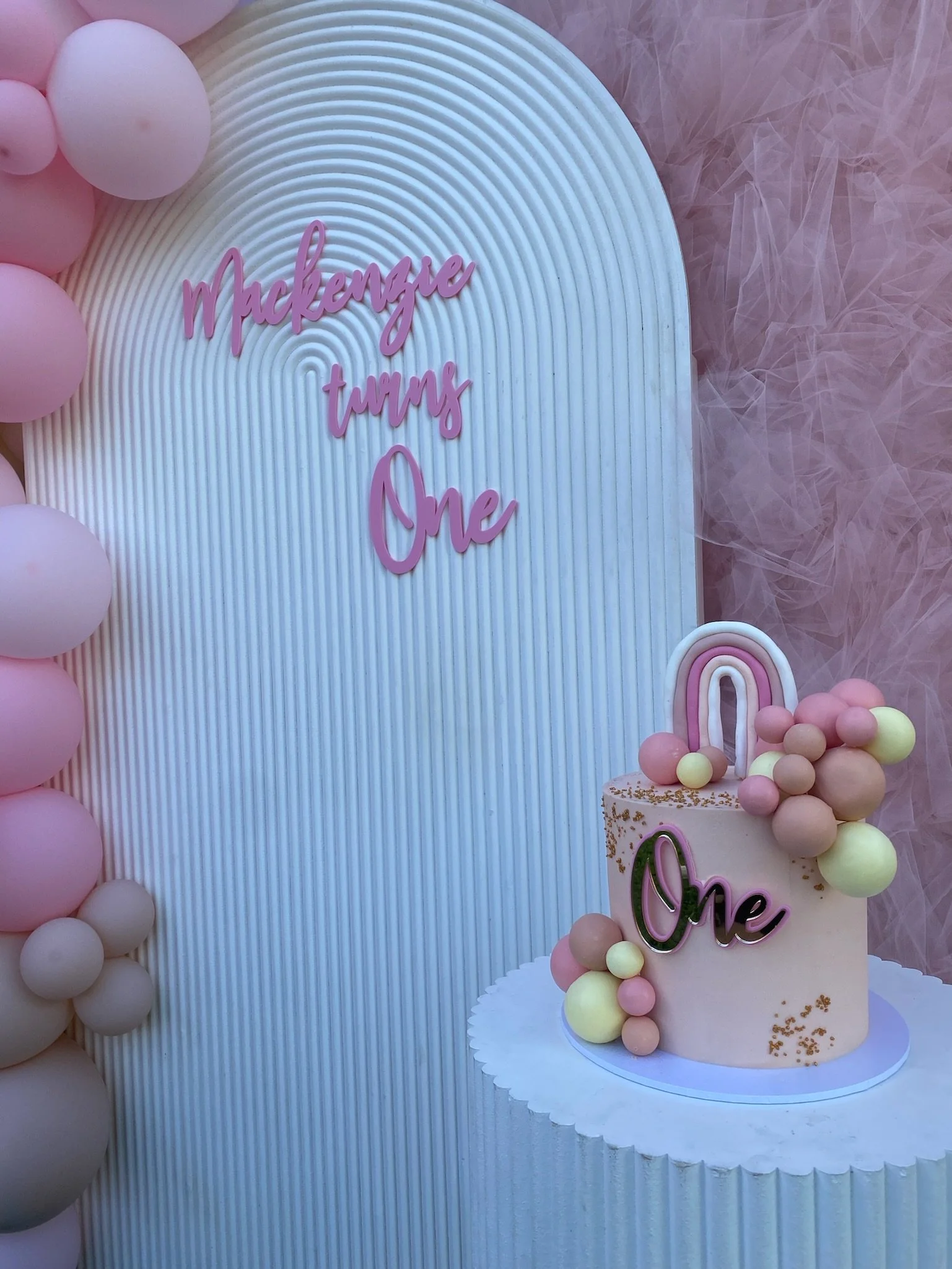 A birthday display with a pastel pink and yellow theme, featuring a cake decorated with a rainbow topper and the words "One" in pink, alongside a backdrop with the phrase "Mackenzie turns One".