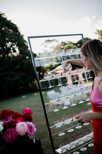 A woman in a pink dress reaching into a glass display of champagne glasses outdoors, with a bouquet of pink and red roses nearby and trees in the background.