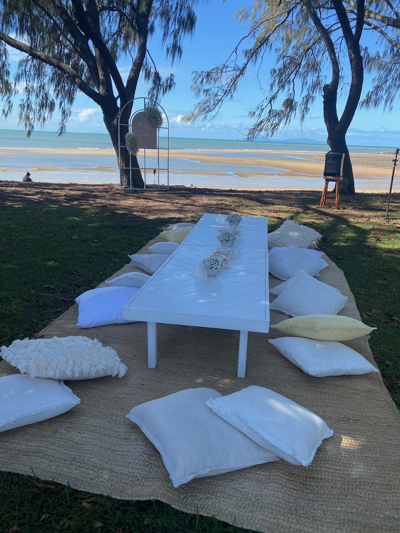 A beachside setup with a low white table surrounded by white and yellow pillows on a woven rug, with a sandy beach and ocean in the background under trees.