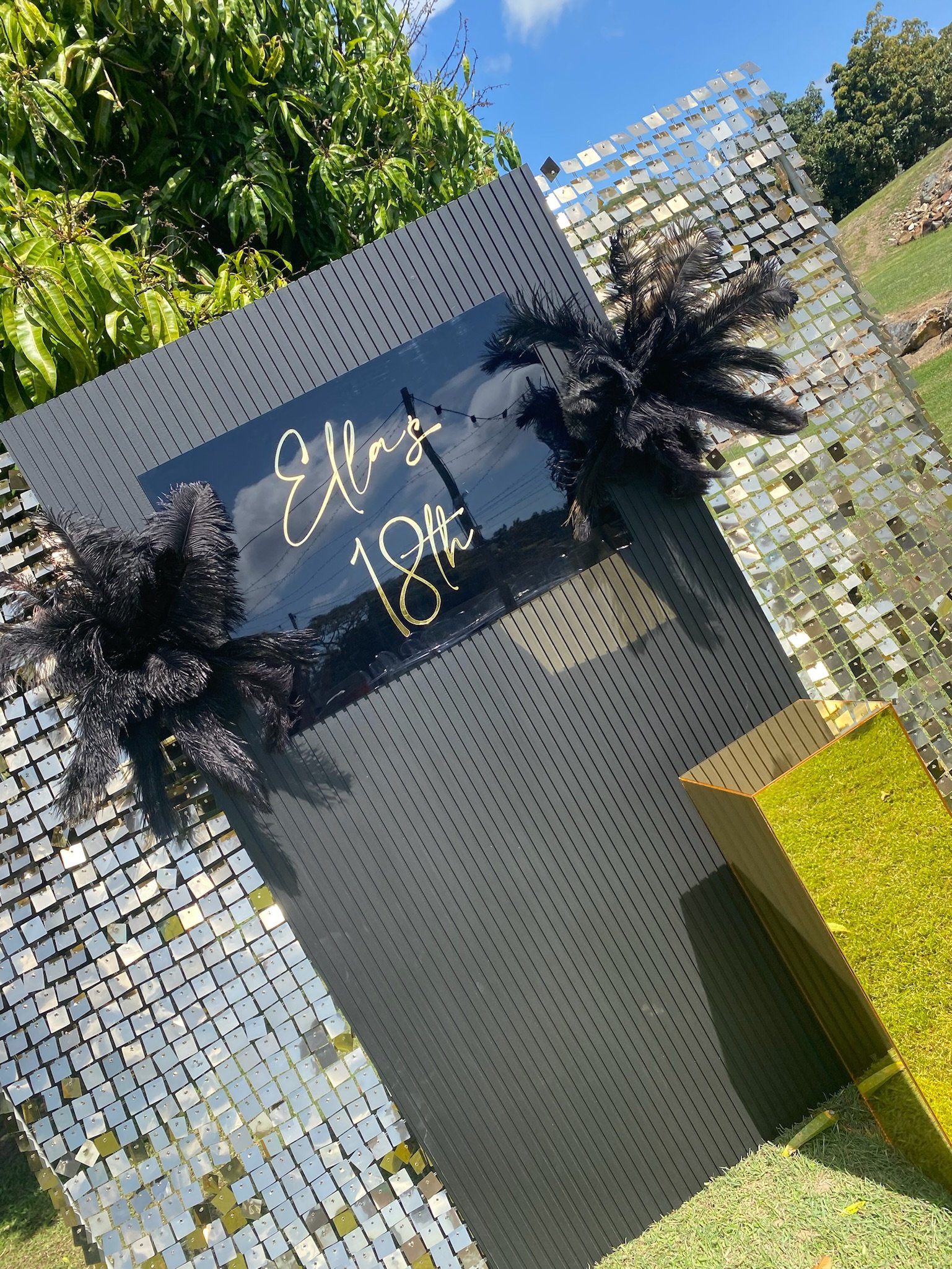 A decorative outdoor display with a reflective black sign reading 'Elias 18th' in yellow script, adorned with black feathered decorations, set against a backdrop of greenery and a blue sky.