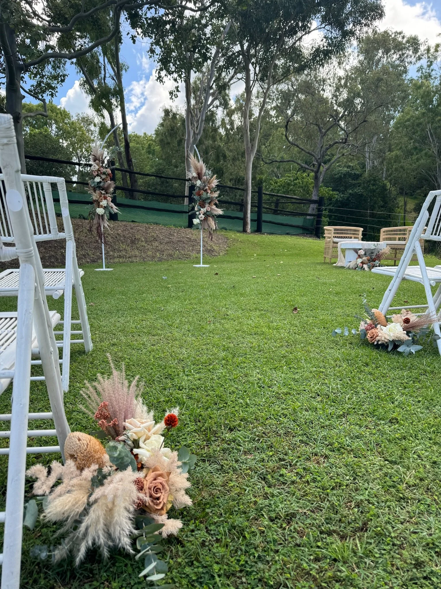 Outdoor wedding ceremony setup with white chairs, floral arrangements, and an arch in a grassy area surrounded by trees.