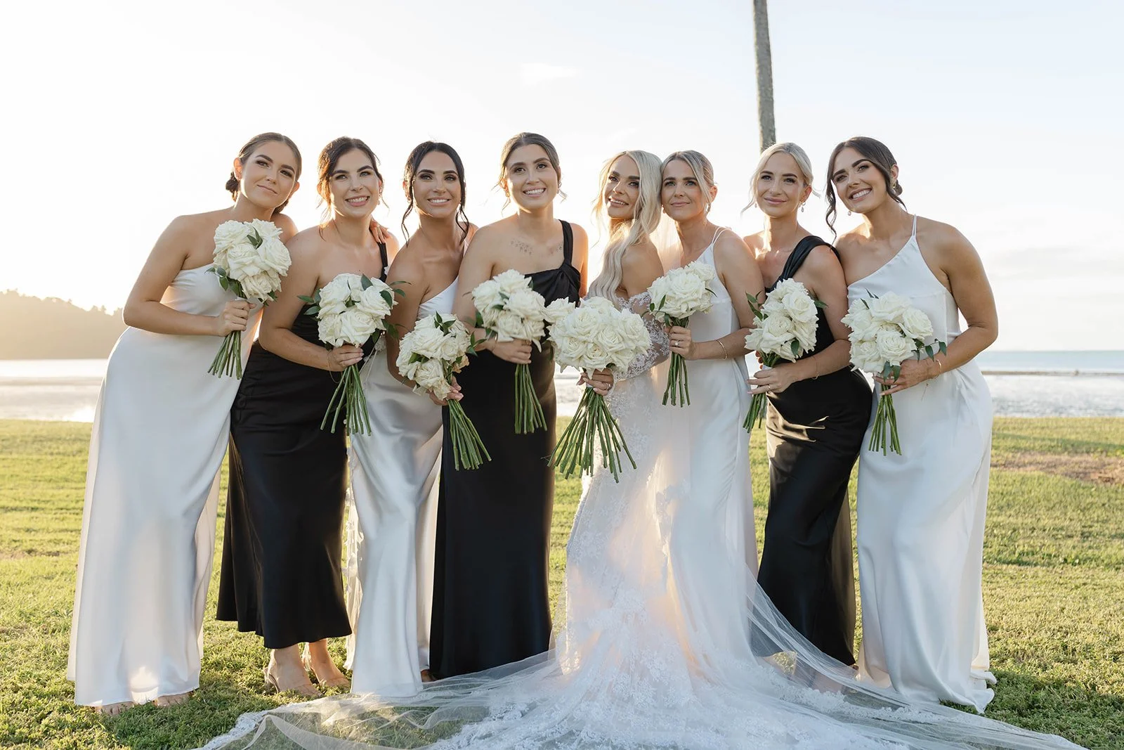 Bride and bridesmaids standing outdoors on grass at sunset, holding bouquets of white roses, smiling at camera.