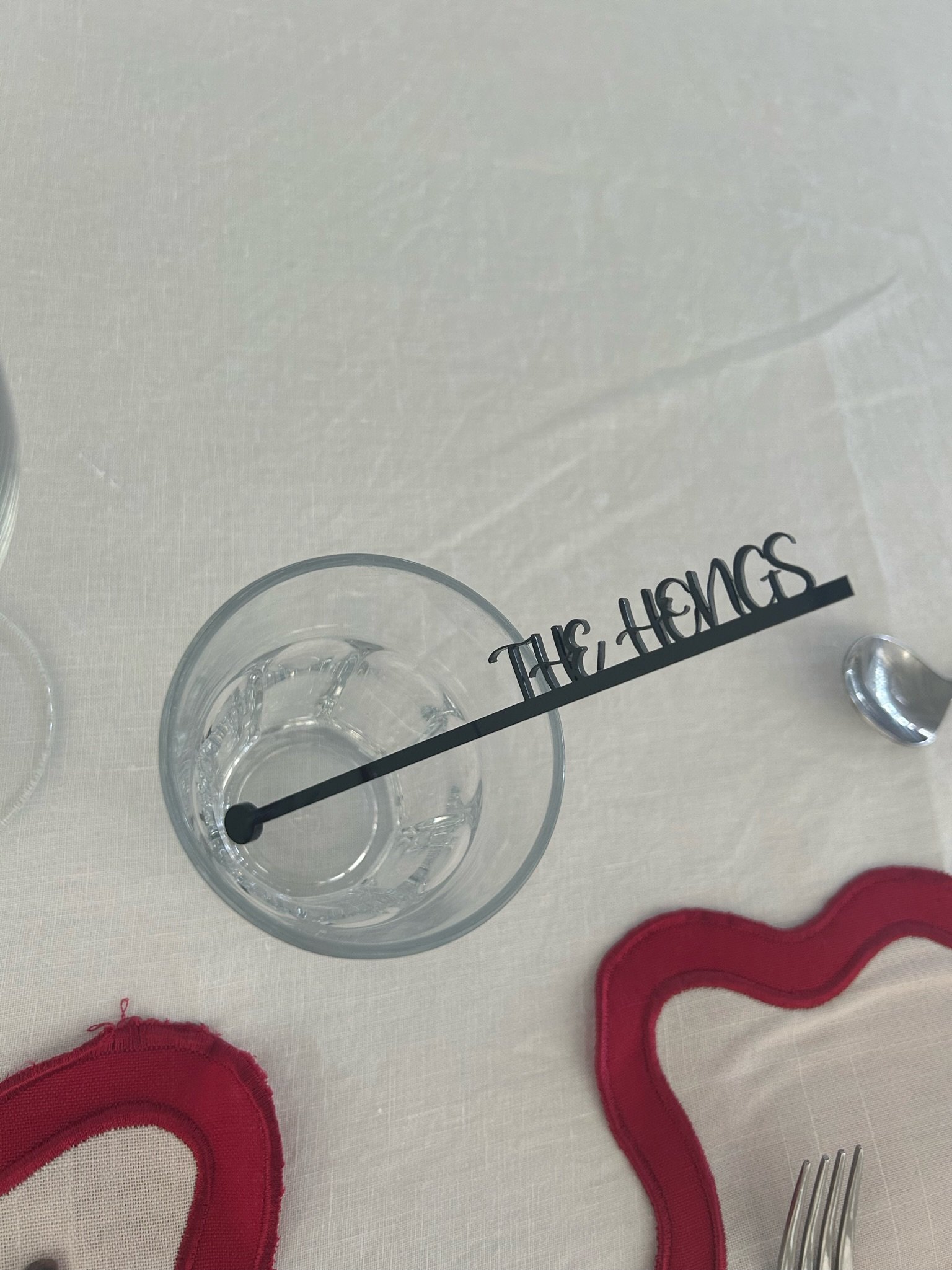 A table with a glass of water, red heart-shaped fabric decorations, and a table marker reading 'The Hangs' with a shadow, on a white tablecloth.