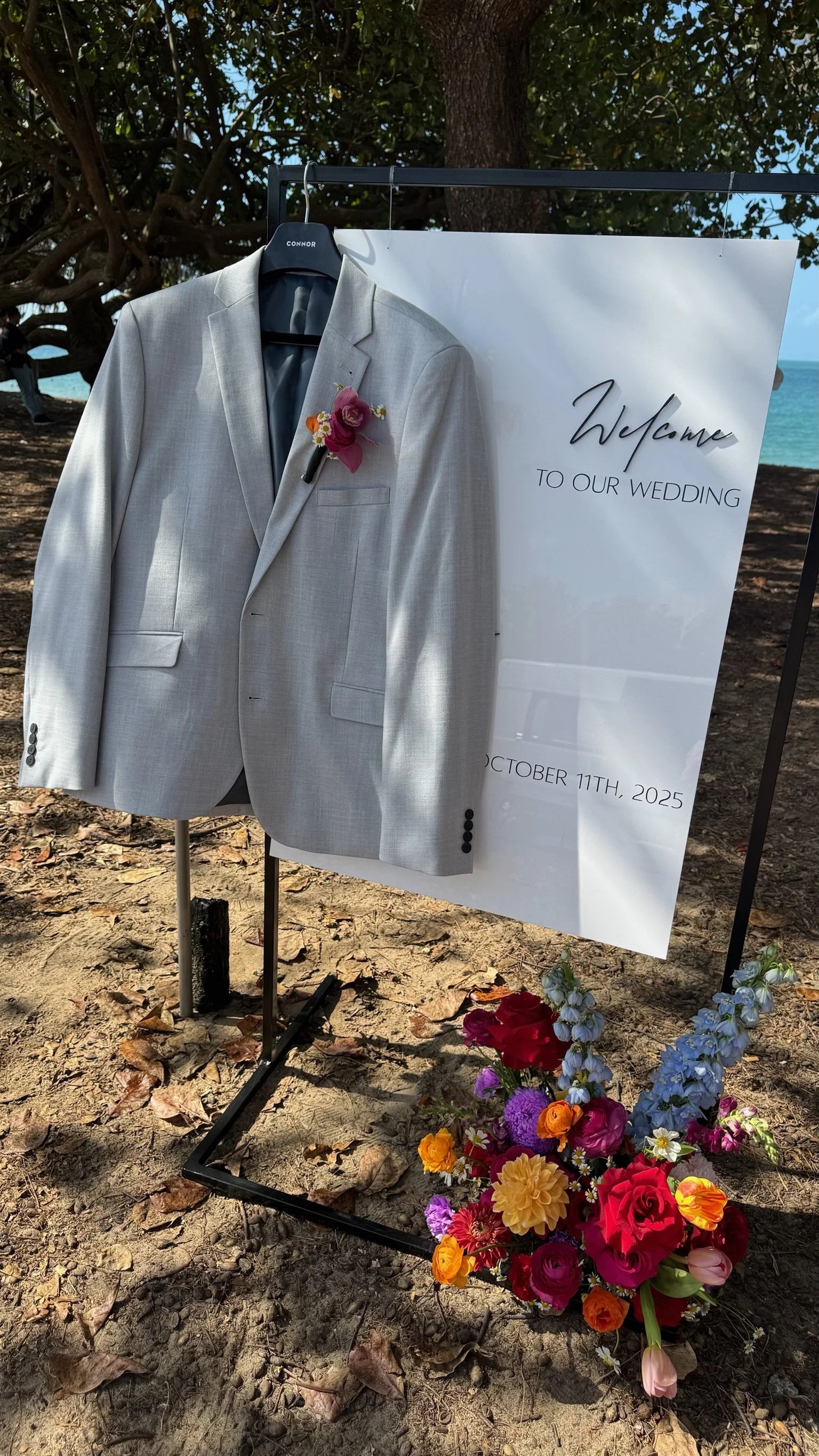 A light grey suit jacket hung on a black hanger with the name 'CONNOR' on it, hanging on a frame near a sign that reads 'Welcome to our wedding' with the date October 11th, 2025, in front of trees and a glimpse of the ocean in the background. There's
