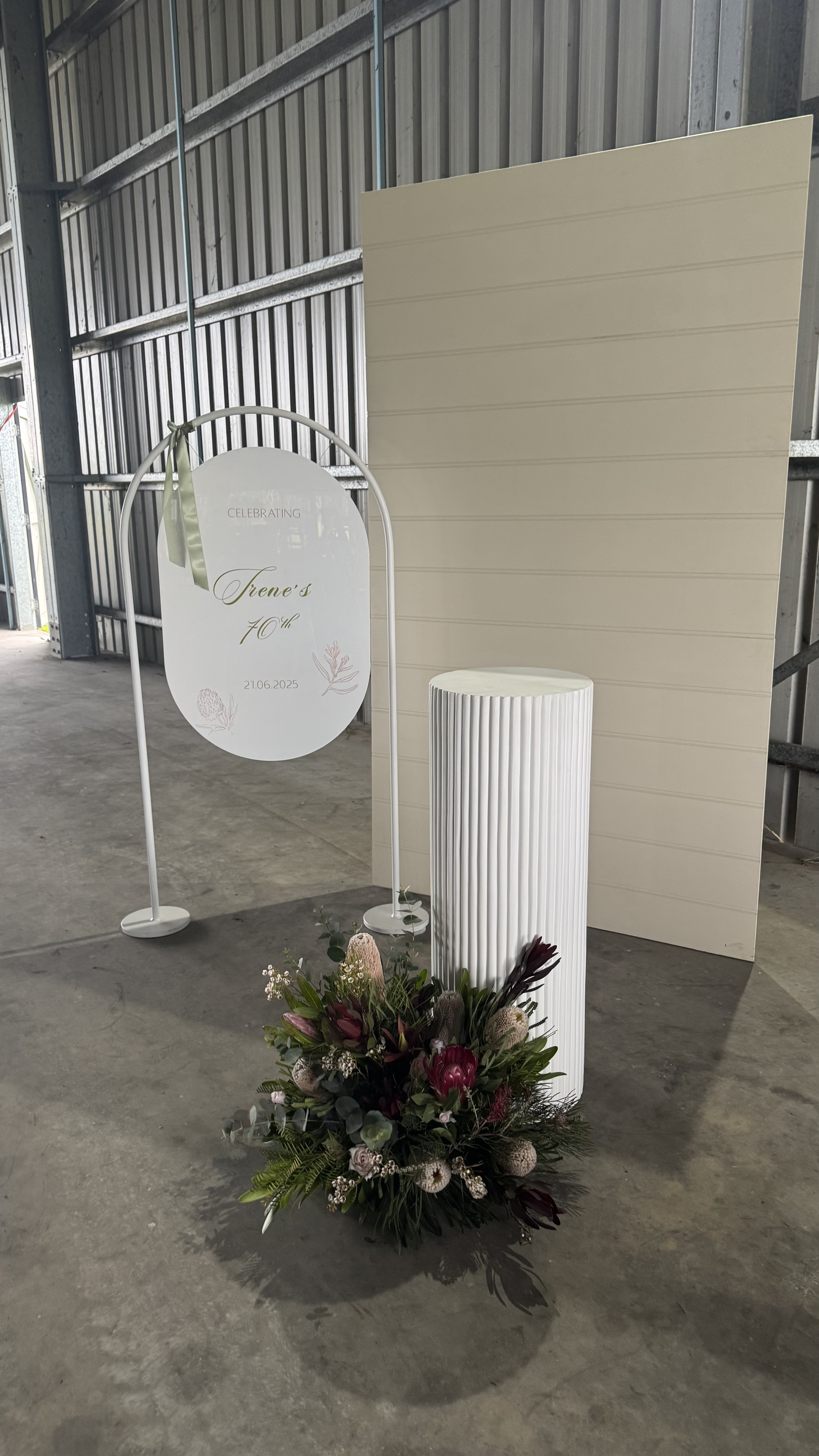 A decorative setup for a 40th celebration featuring a white oval sign with pastel-colored lettering, a floral arrangement on the ground, a white cylindrical pillar, and a beige wall panel in the background, inside a metal-walled building.