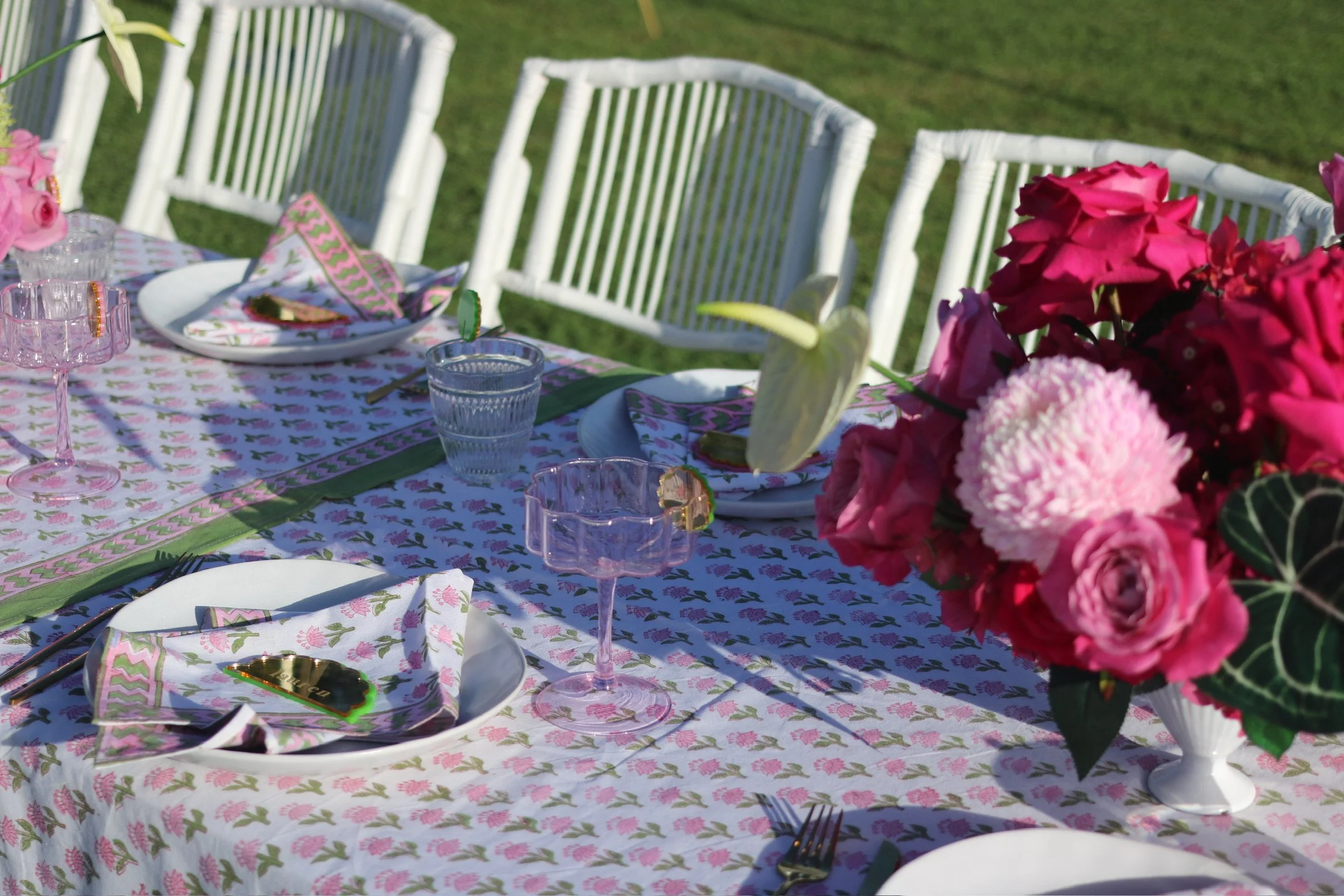 Table set for an outdoor gathering with pink floral tablecloth, pink and clear glassware, white plates with patterned napkins, and a large bouquet of pink and white flowers.