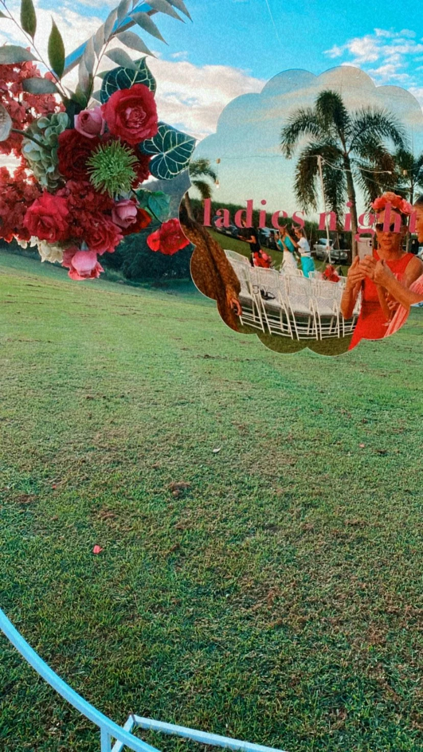 Part of a floral photo booth mirror with a reflection of women at an outdoor event, including palm trees, chairs, and people, with the phrase "ladies nigh" visible in the reflection.