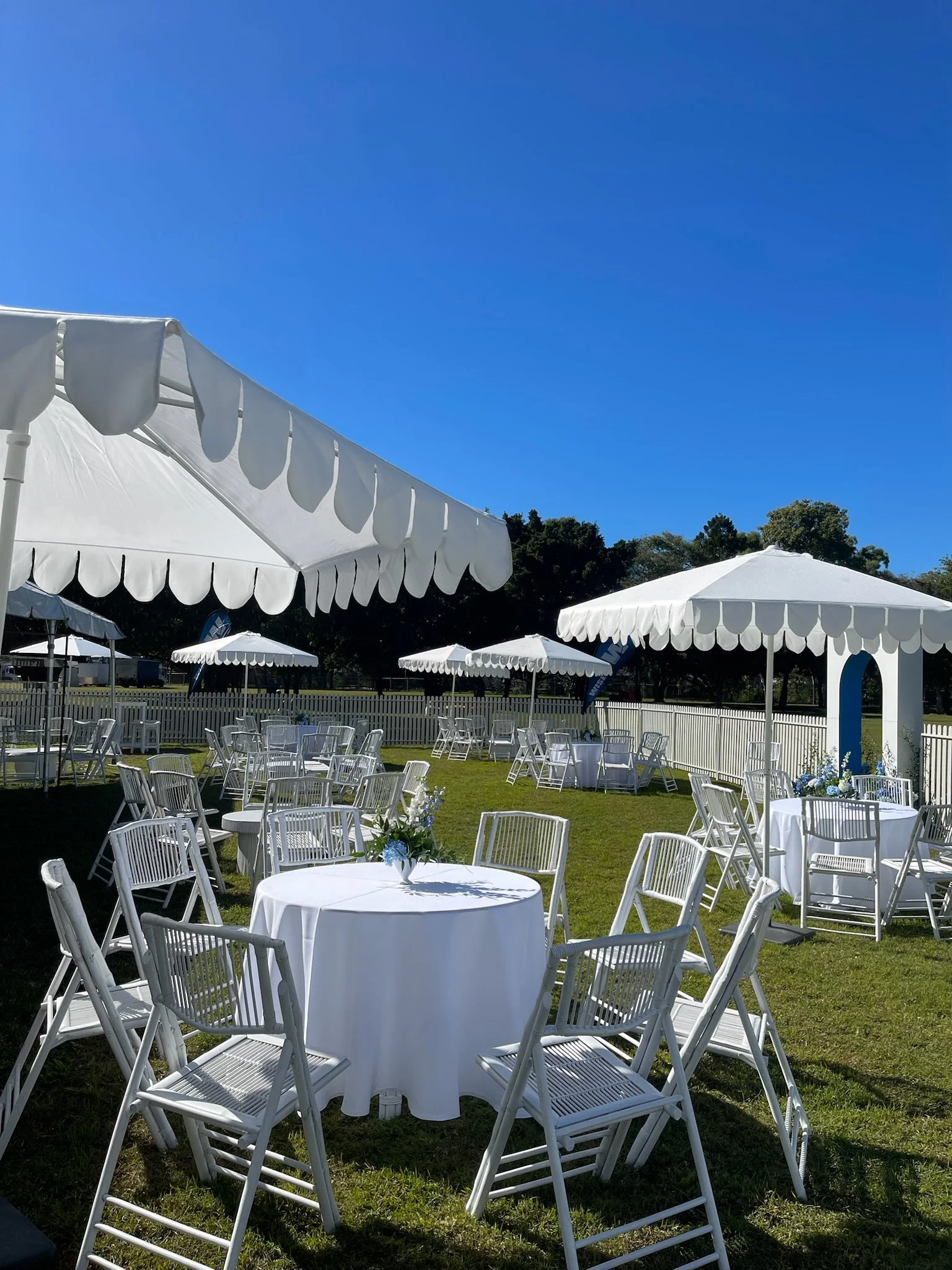 Outdoor setup with white tables, chairs, and large white umbrellas under clear blue sky