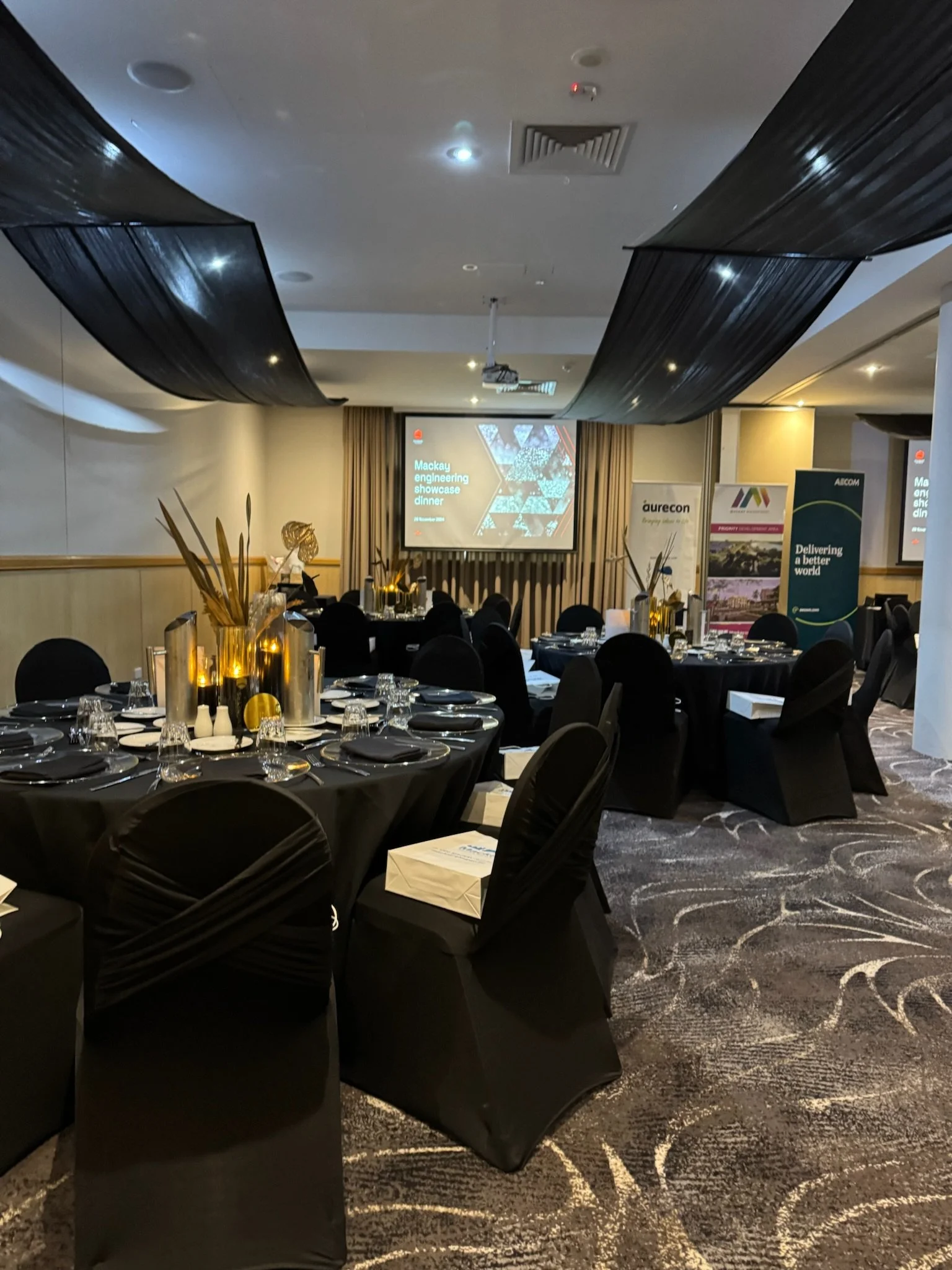 A banquet room set up for an event with round tables covered in black tablecloths, decorated with gold vases and tableware, and a projector screen displaying 'Mackay engineering showcase dinner' in the background.