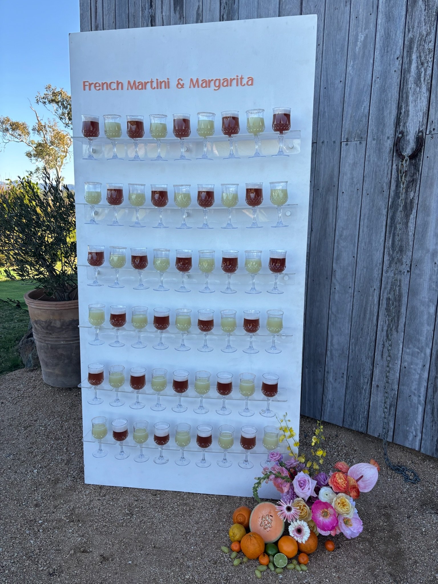 Board titled "French Martini & Margarita" with multiple rows of small glasses filled with red and green drinks, arranged on shelves. There is a floral arrangement with pink, purple, orange flowers, and fruits including oranges and limes at the base o