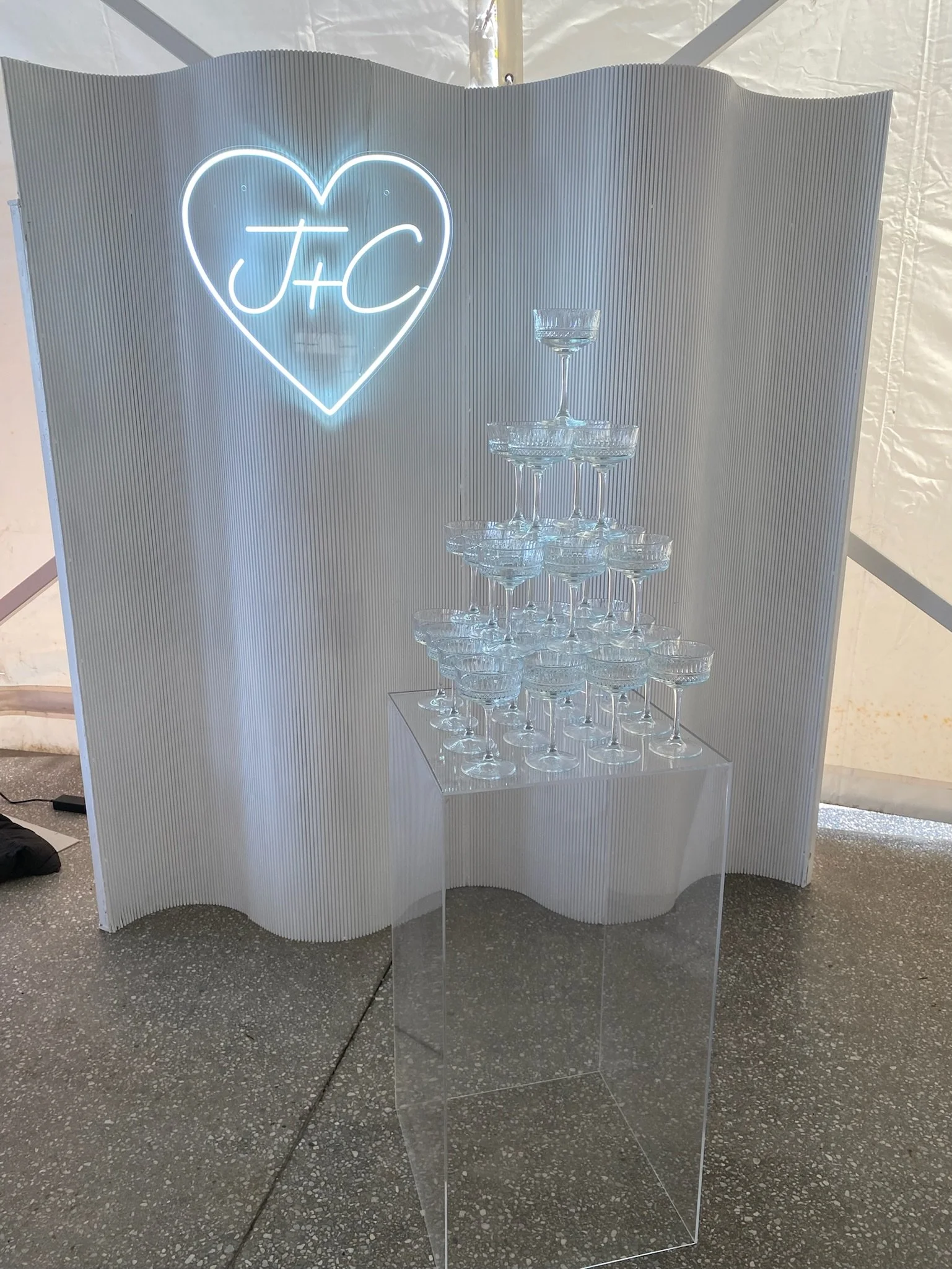 A display with a neon heart sign with the initials J+C inside, and a champagne tower made of stacked coupe glasses on a clear rectangular stand, set against a white textured backdrop.