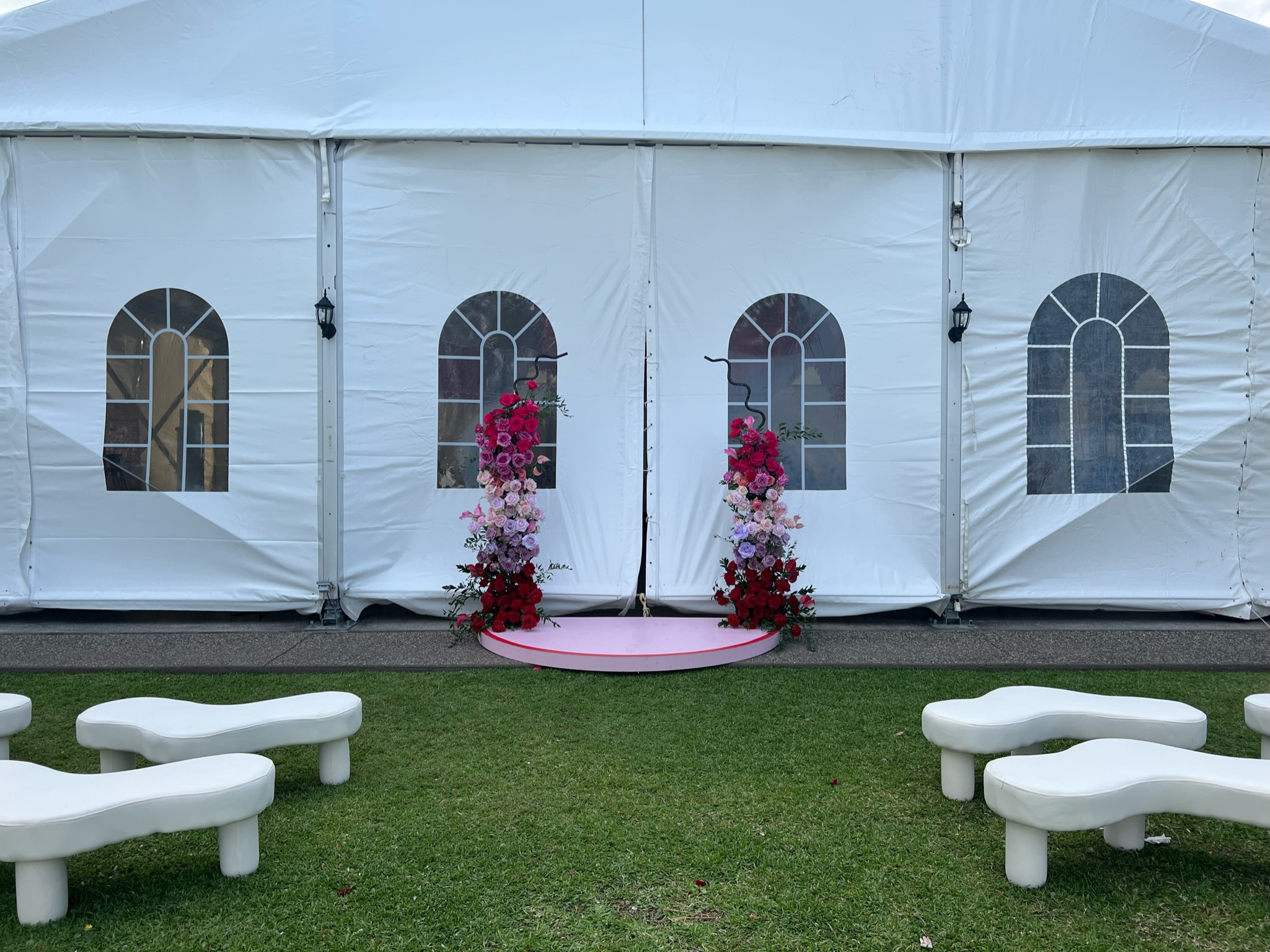A white event tent decorated with a flower arch on a pink platform in front of the entrance. The tent has arched windows and is set on a grassy area with seating nearby.