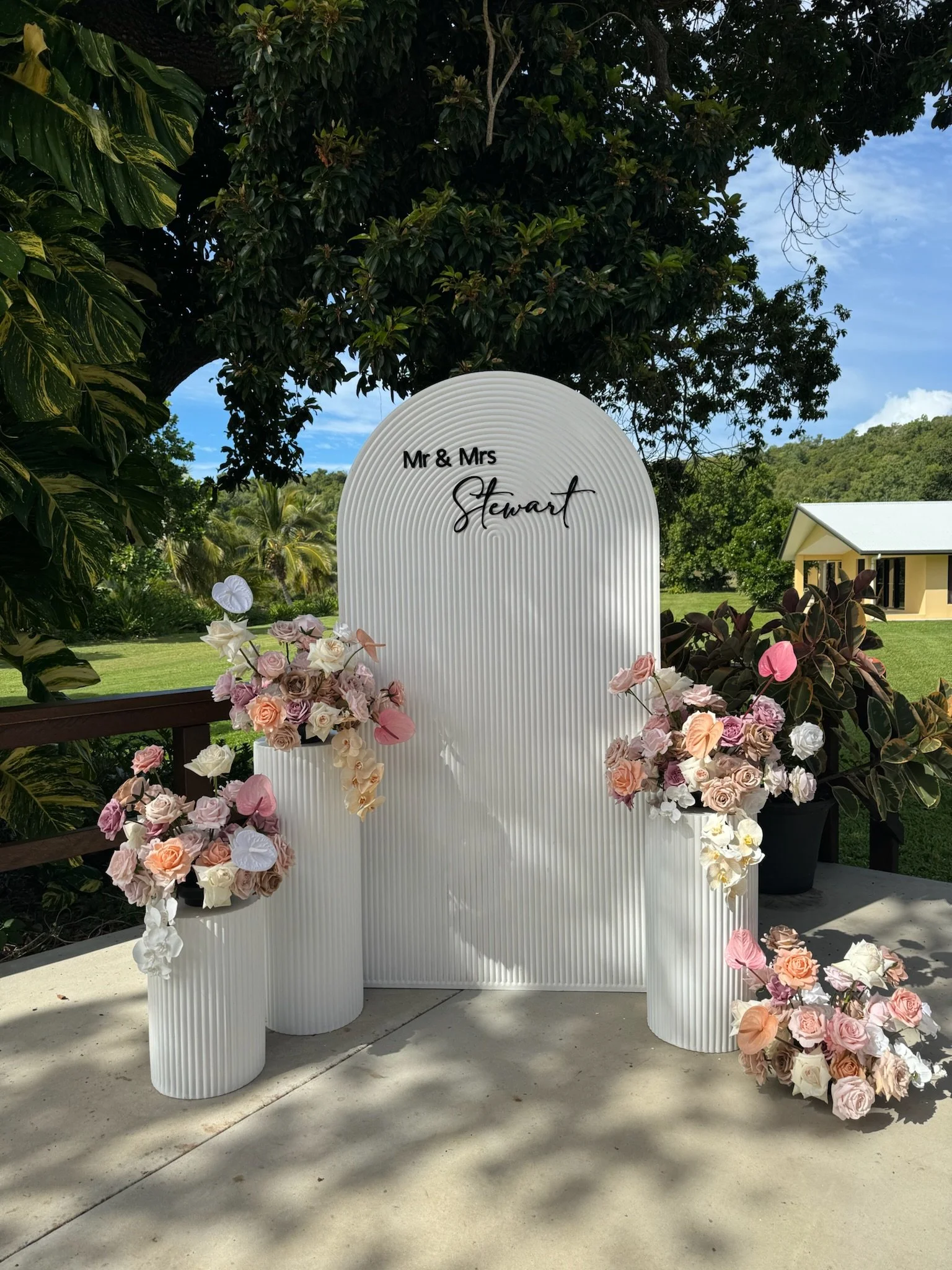 Outdoor wedding or event backdrop with a white ribbed arch and floral arrangements in pastel colors on white vases, set against greenery and a blue sky.