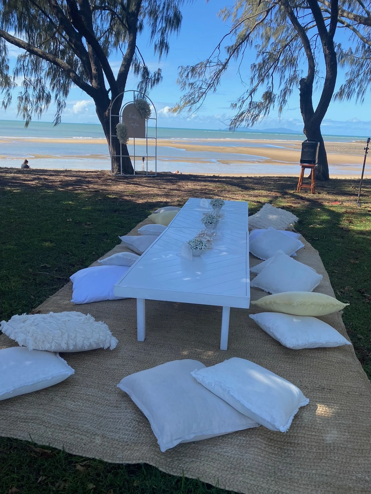 Beachside setup with a white table on a woven mat surrounded by white and cream pillows, overlooking a sandy beach and ocean with blue sky and trees nearby.