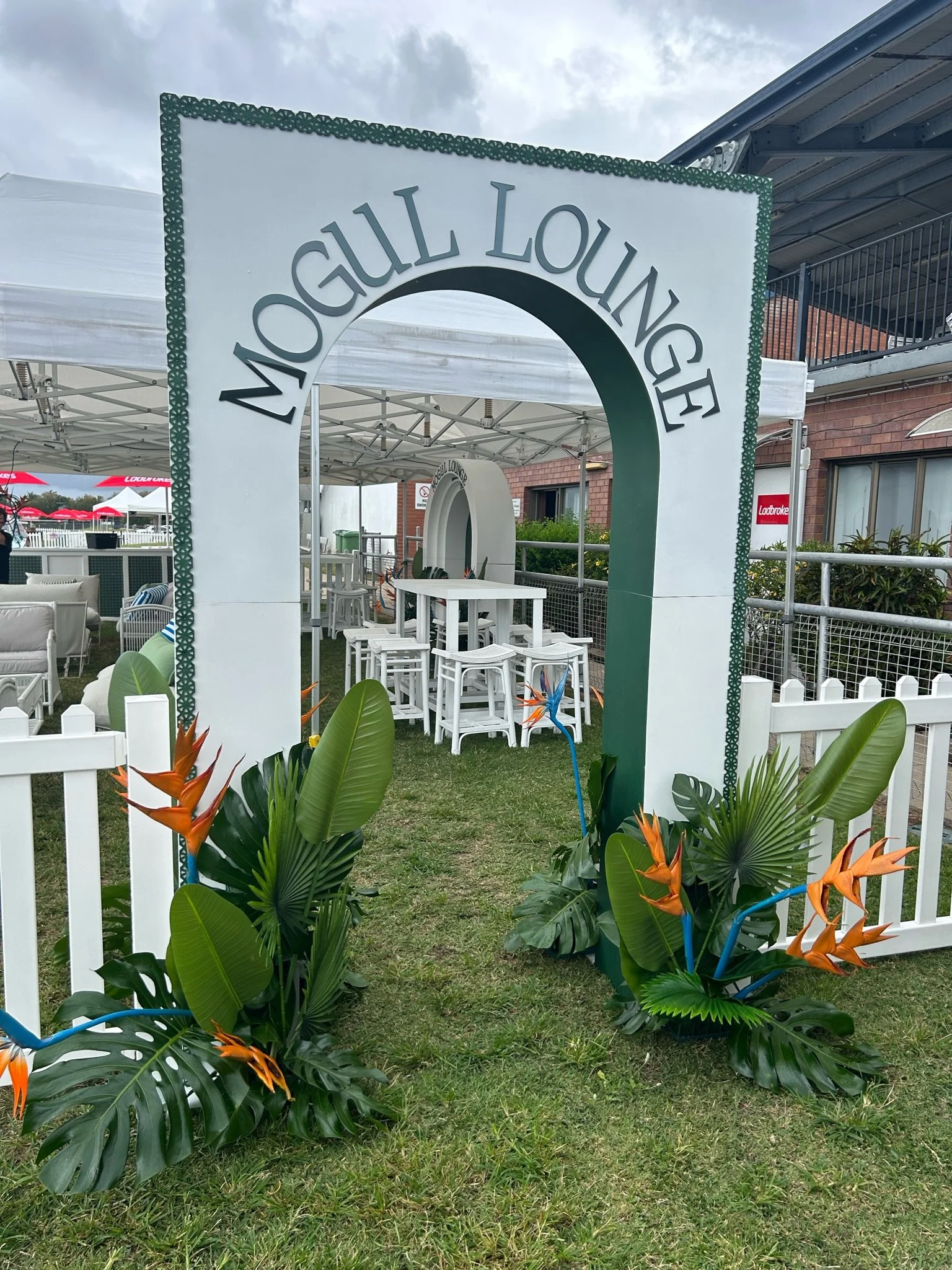 Entrance to a venue called 'Mogul Lounge' with a white sign archway, tropical plants with orange flowers at the base, white fence, outdoor seating, and tents in the background under cloudy skies.