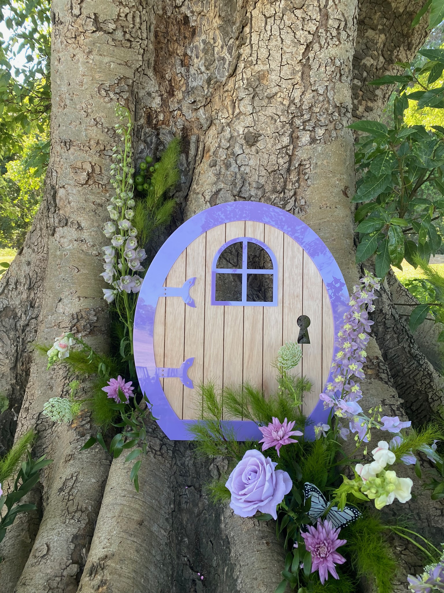 A whimsical door with purple trim and a small arched window, set against the trunk of a large tree, decorated with flowers and greenery.
