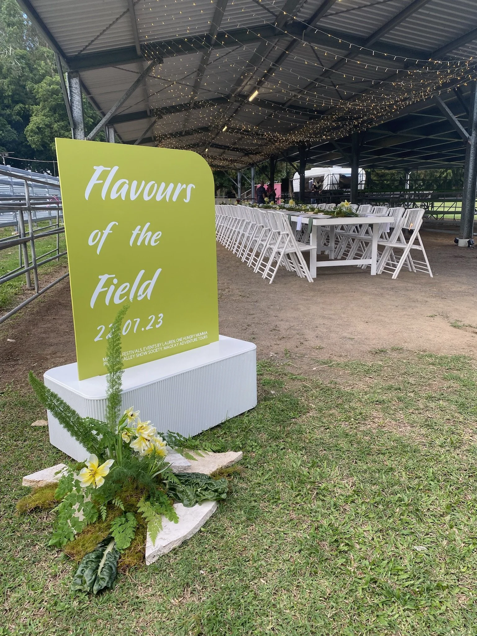 An outdoor event setup with a large green sign reading 'Flavours of the Field 22.07.23' near a flower arrangement, with long white tables and chairs under a metal roof decorated with string lights.