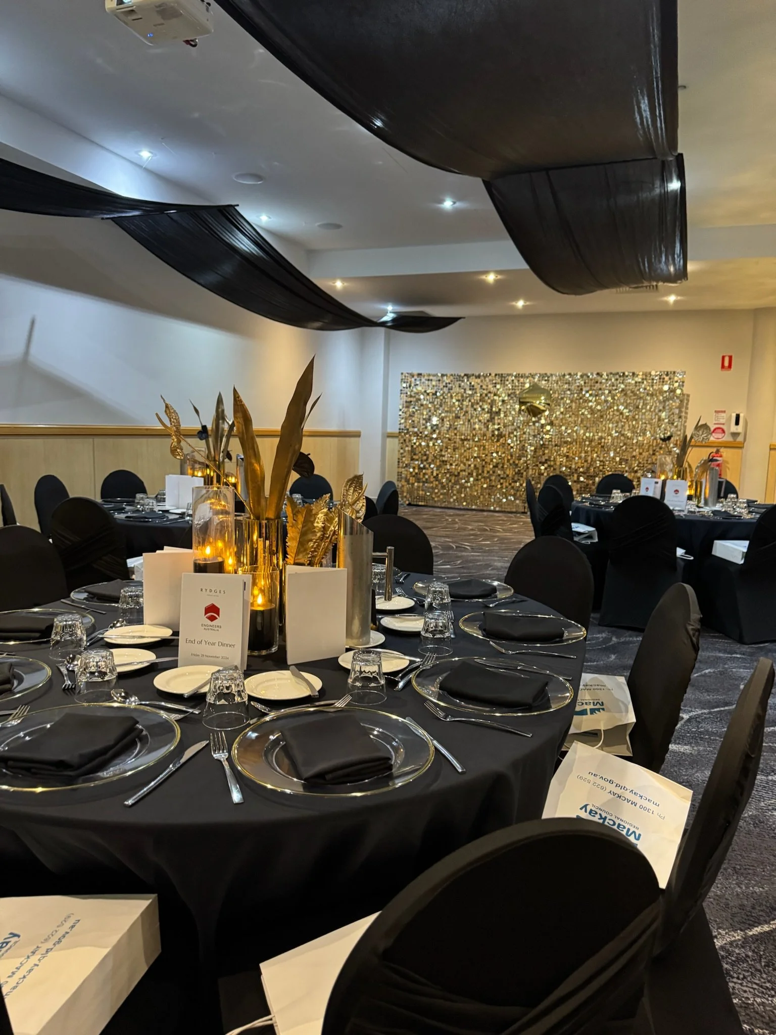 A banquet hall decorated with black tablecloths, black chairs, gold accents, and black drapes hanging from the ceiling, set for an event with plates, glasses, utensils, and centerpieces.