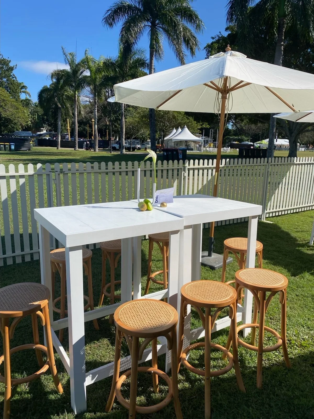 An outdoor scene featuring a white table with a white umbrella, surrounded by wooden stools on green grass, with palm trees and tents in the background under a blue sky.