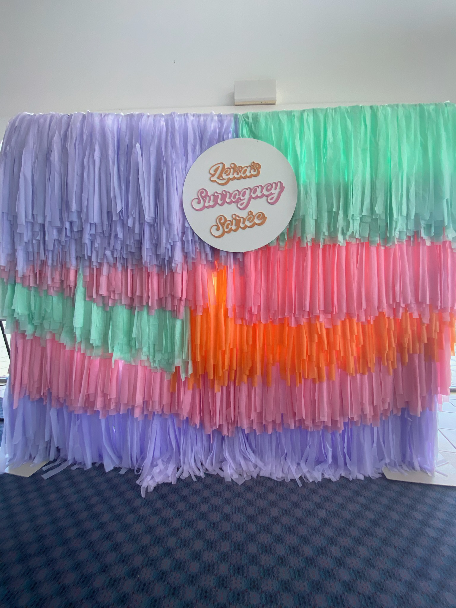 Colorful photo booth backdrop with pastel tissue paper fringe in purple, pink, green, and orange, and a round sign that reads 'Leisa's Surrogacy Soirée' in cursive lettering.