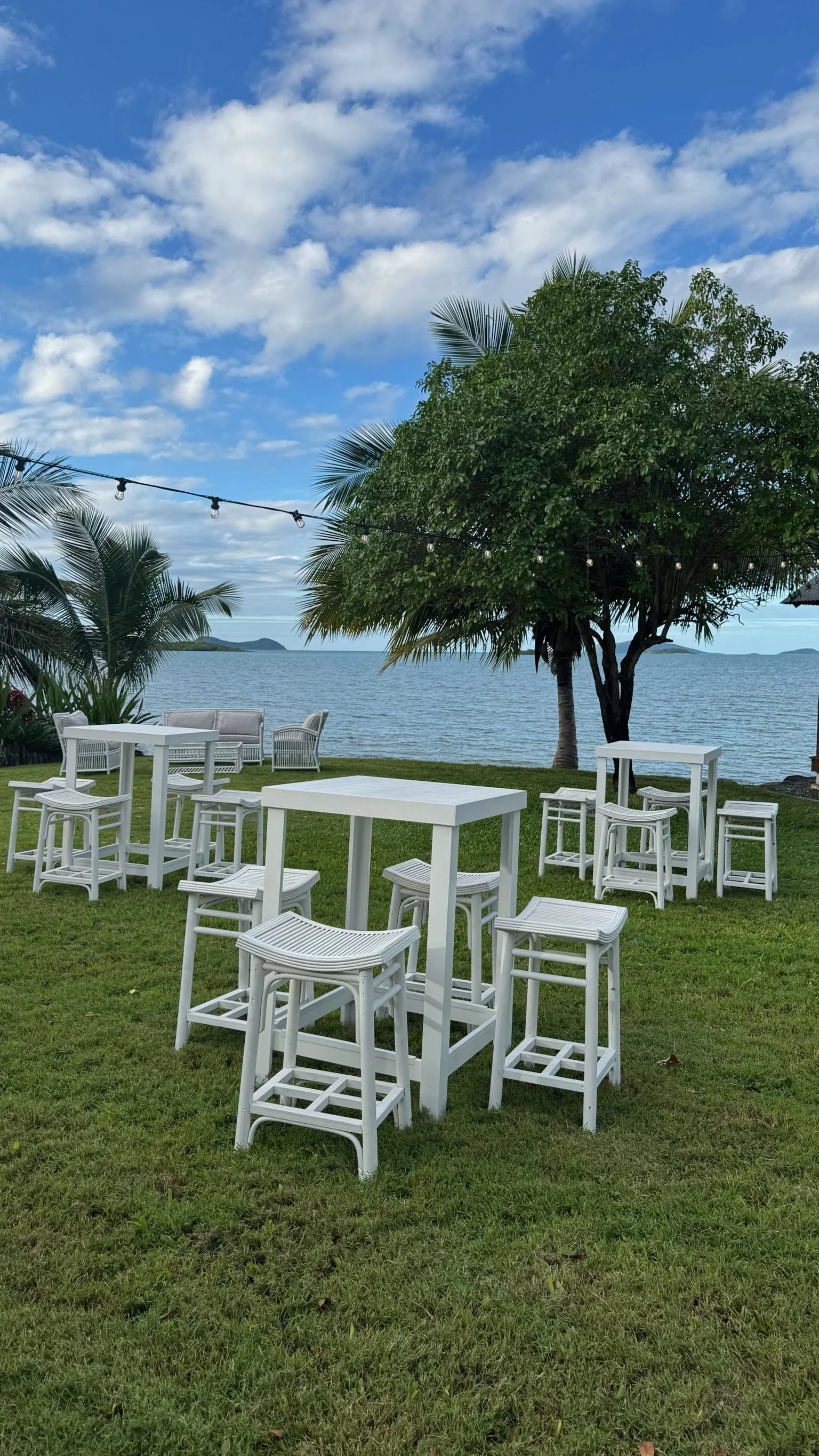 Outdoor seating area with white tables and chairs on a grassy lawn near a body of water, with trees and a cloudy sky in the background.