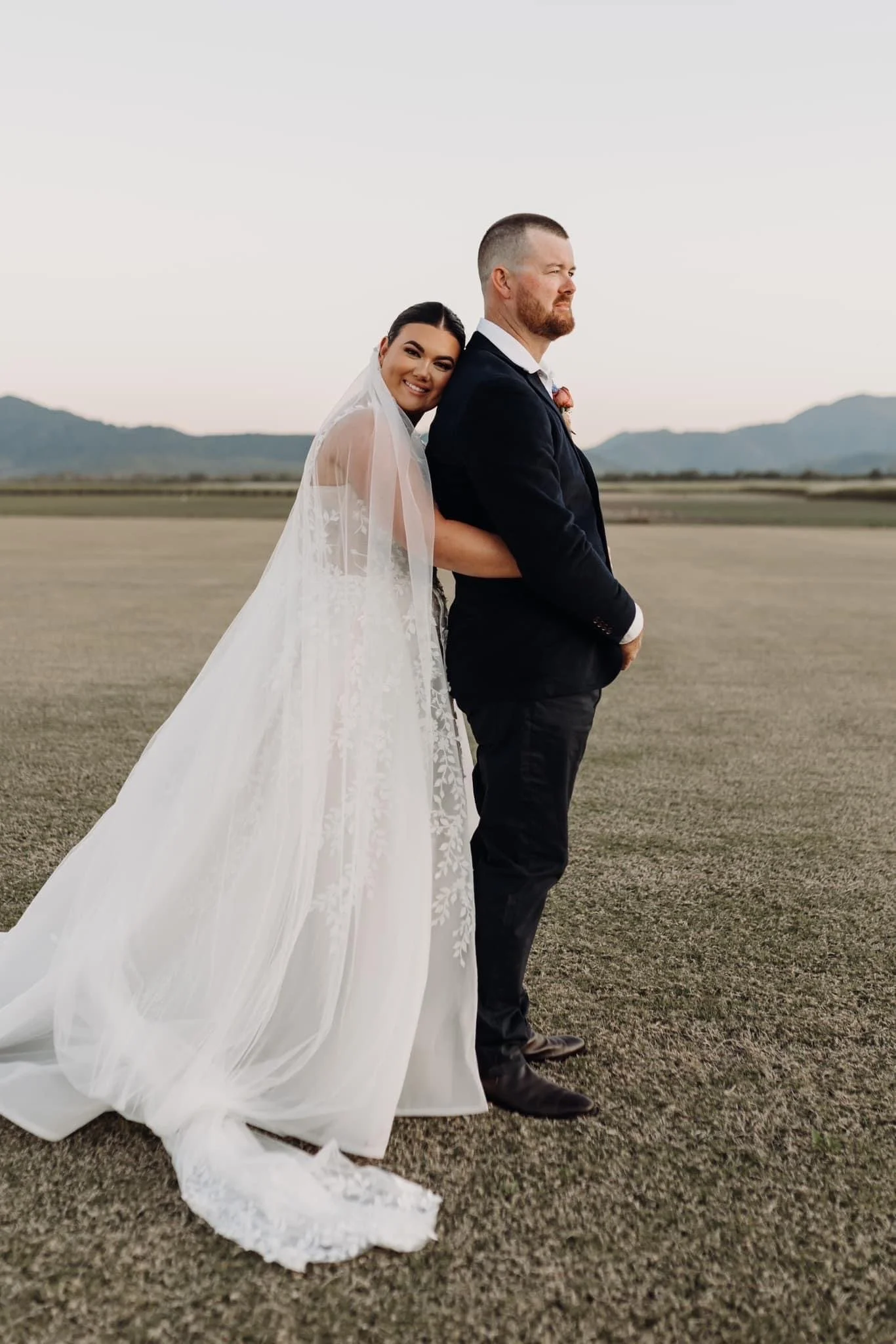 A bride and groom standing outdoors on a grassy field at sunset, with the bride hugging the groom from behind and smiling at the camera. The bride is wearing a white wedding dress with a long train and veil, and the groom is dressed in a black suit w