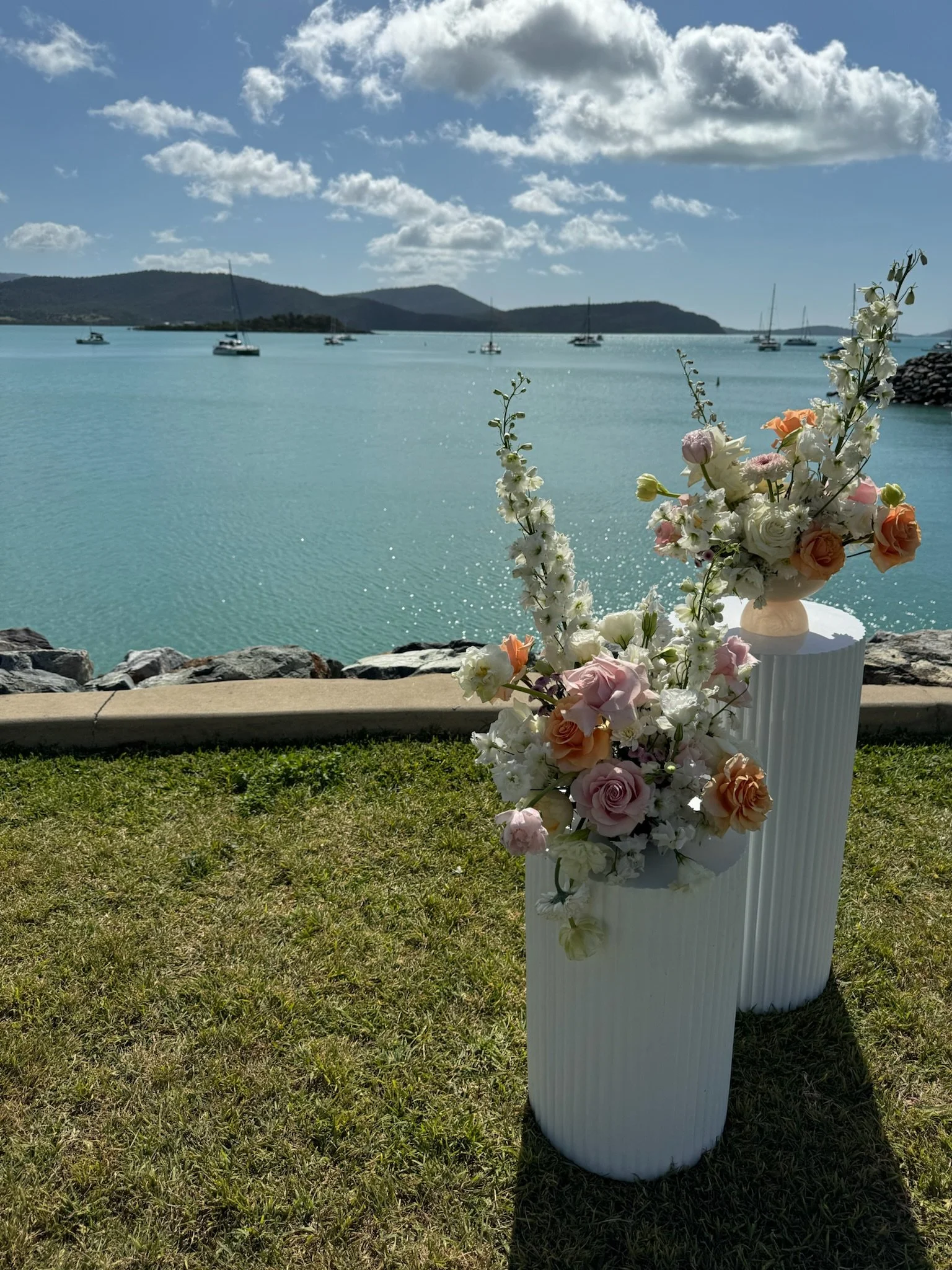 A waterfront scene with two tall white vases filled with pastel-colored flowers, set on grass near a rocky shore, overlooking a calm blue body of water with boats and distant hills under a partly cloudy sky.