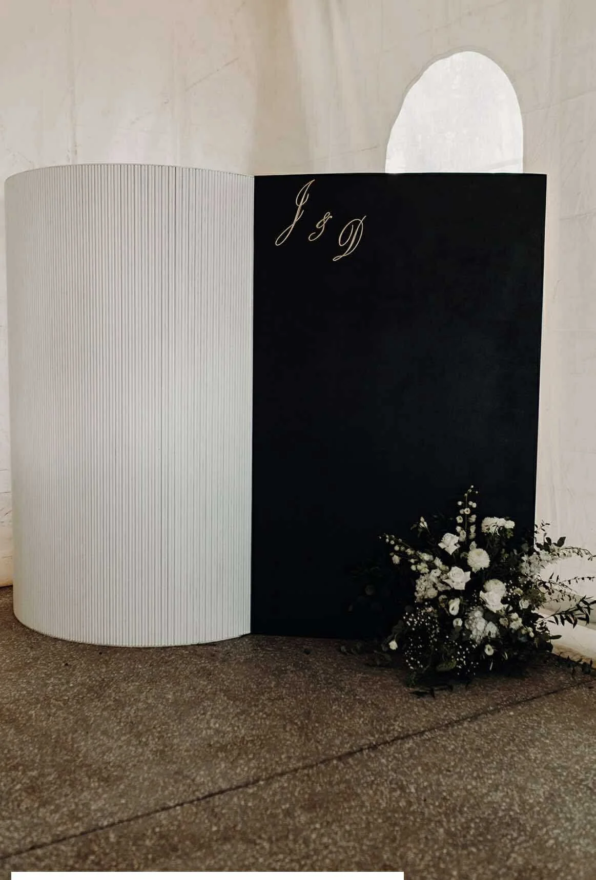 A black and white wedding or event display board with elegant script initials J and D at the top right, and a floral arrangement of white roses and greenery at the base, placed on a textured stone floor against a white wall.