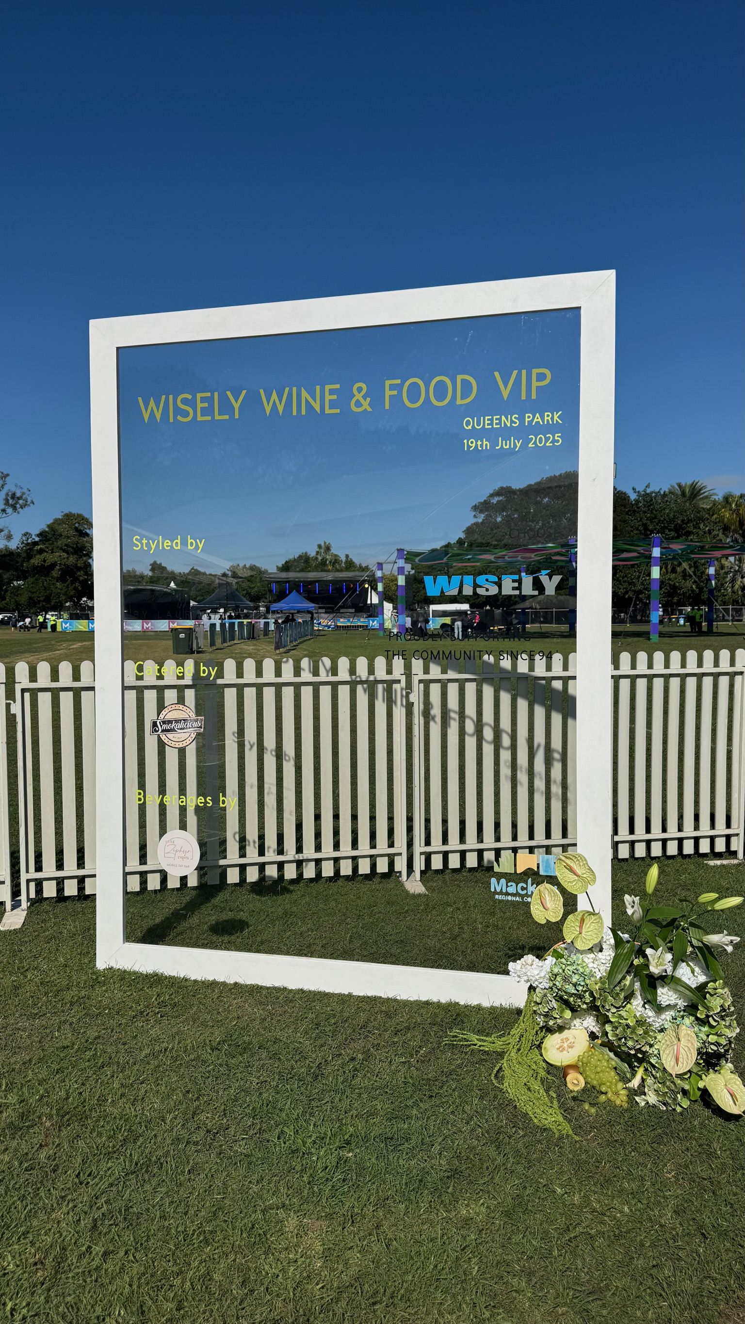 Event signboard for Wisely Wine & Food VIP event at Queens Park on July 19, 2025, with decorations including flowers at the bottom right corner and a fenced outdoor area in the background.