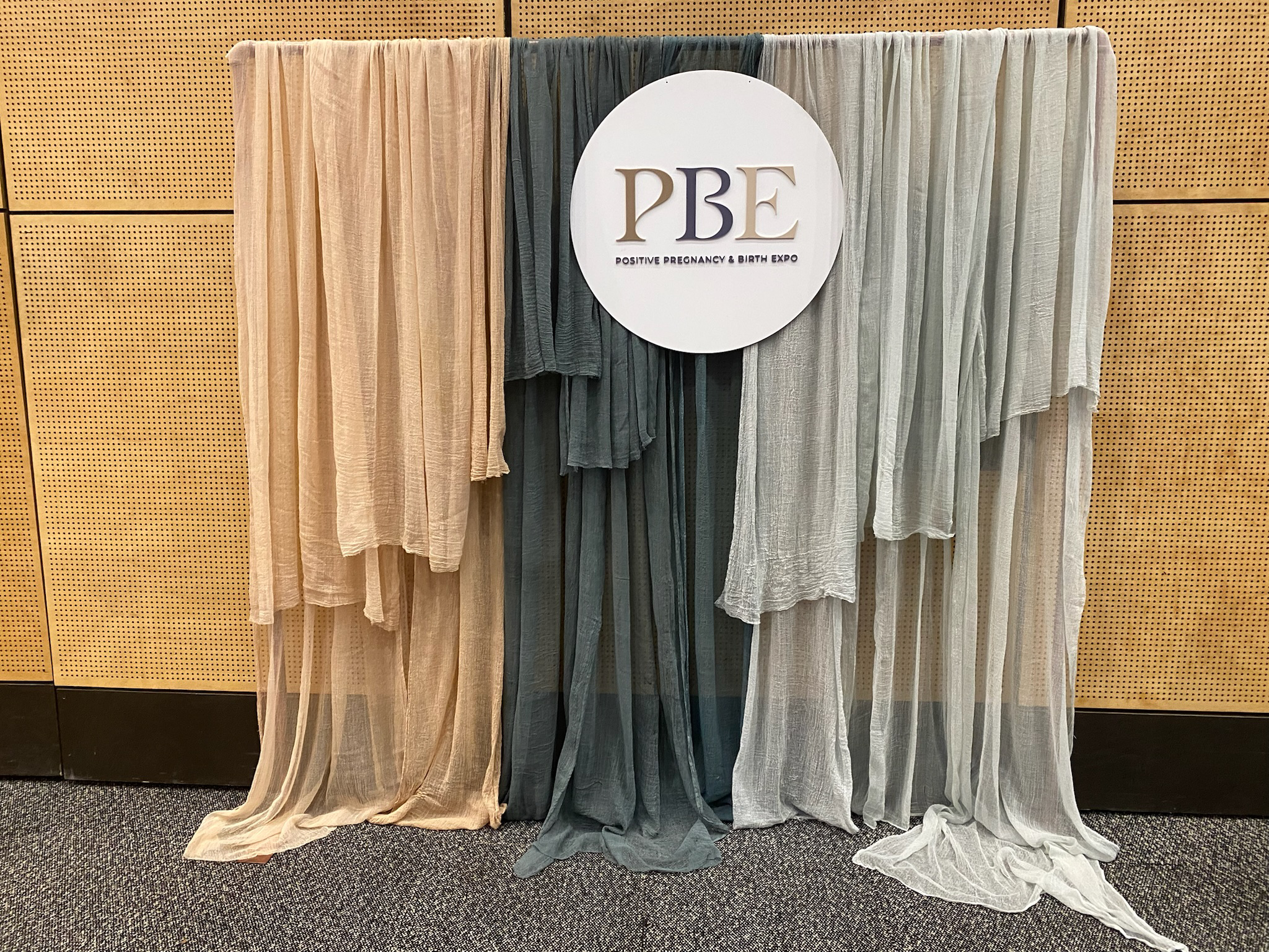 Display of sheer fabrics in neutral colors hanging on a backdrop with a sign that reads 'PBE' and 'Positive Pregnancy & Birth Expo'.