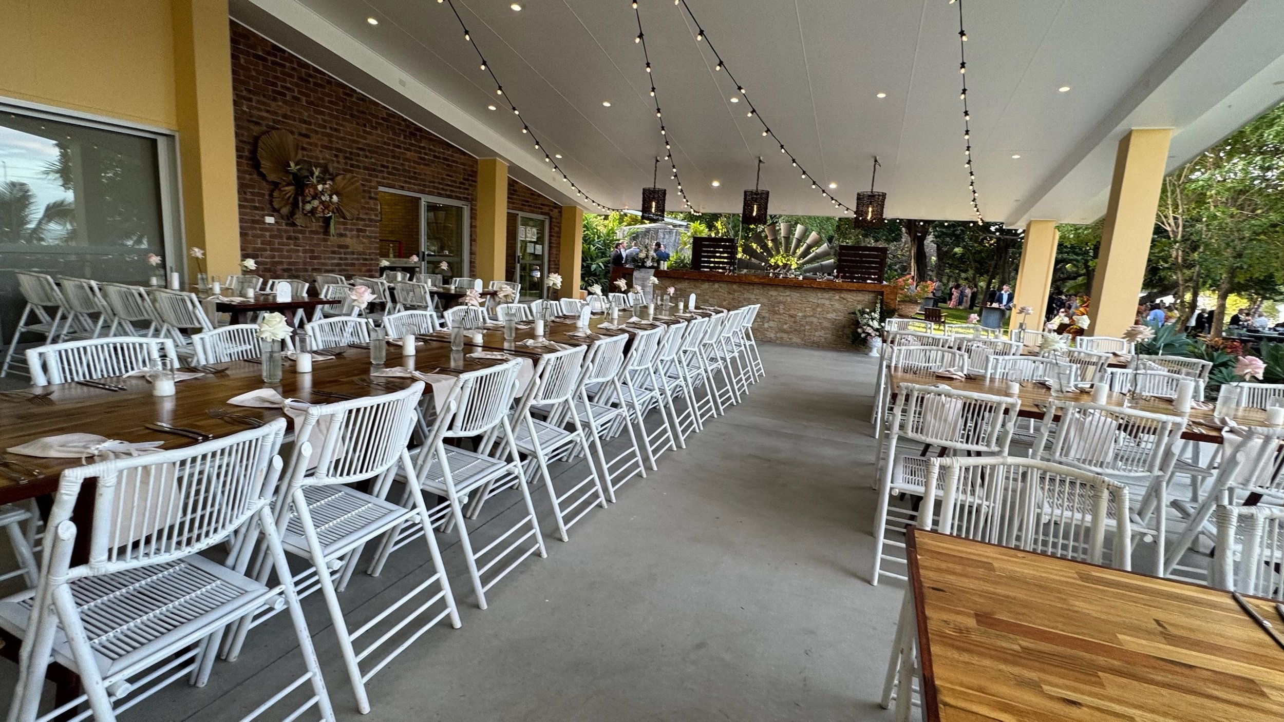 Outdoor event space with long tables set with white flowers, candles, and silverware. String lights hang from the ceiling, and there are trees and people in the background, suggesting a garden party or celebration.