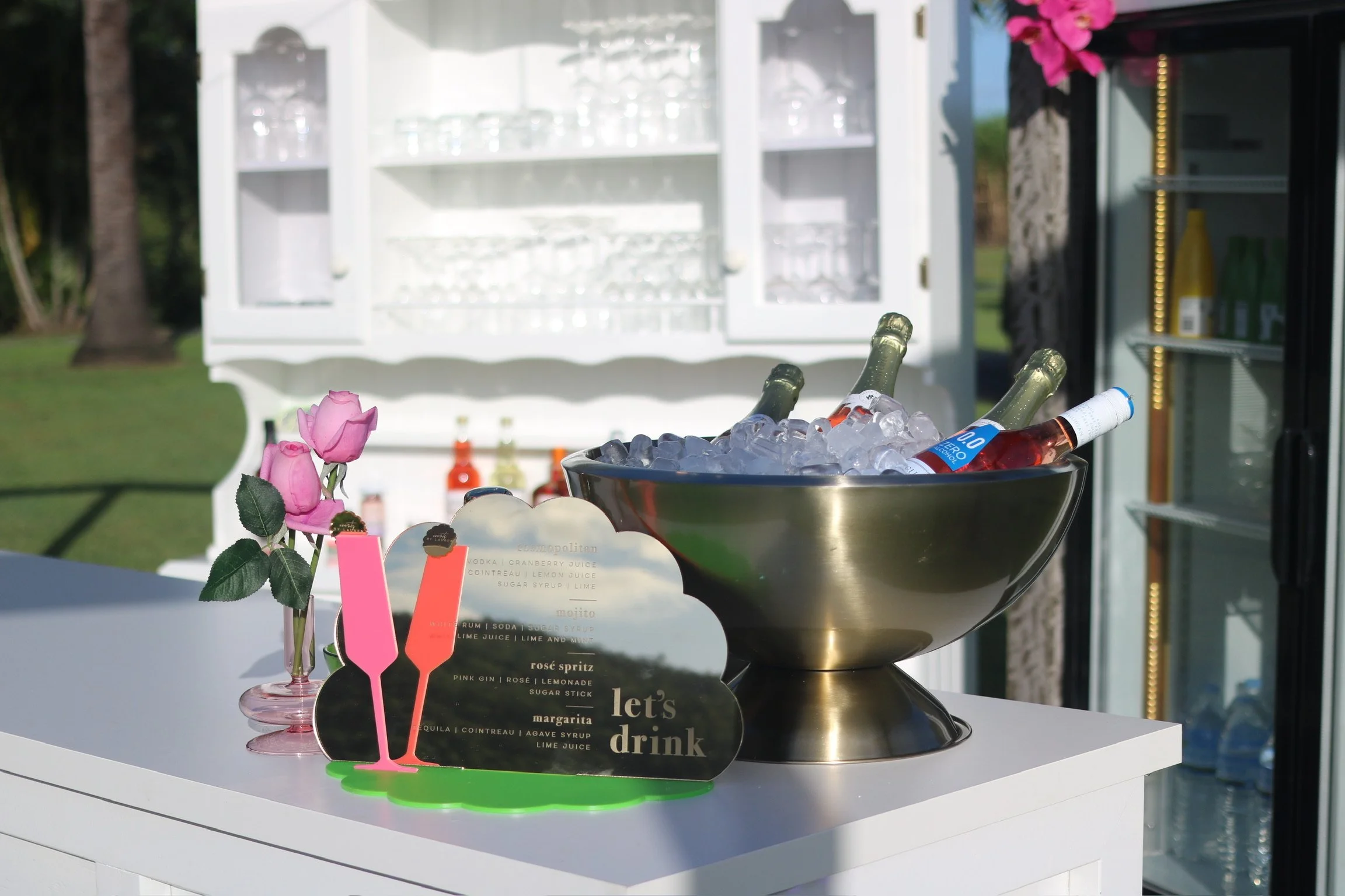 A white outdoor bar cart with glassware and bottles in the background, a metal bowl filled with ice and assorted bottled drinks, a small glass vase with pink roses, and a decorative sign with cocktail menu items that says "let's drink".