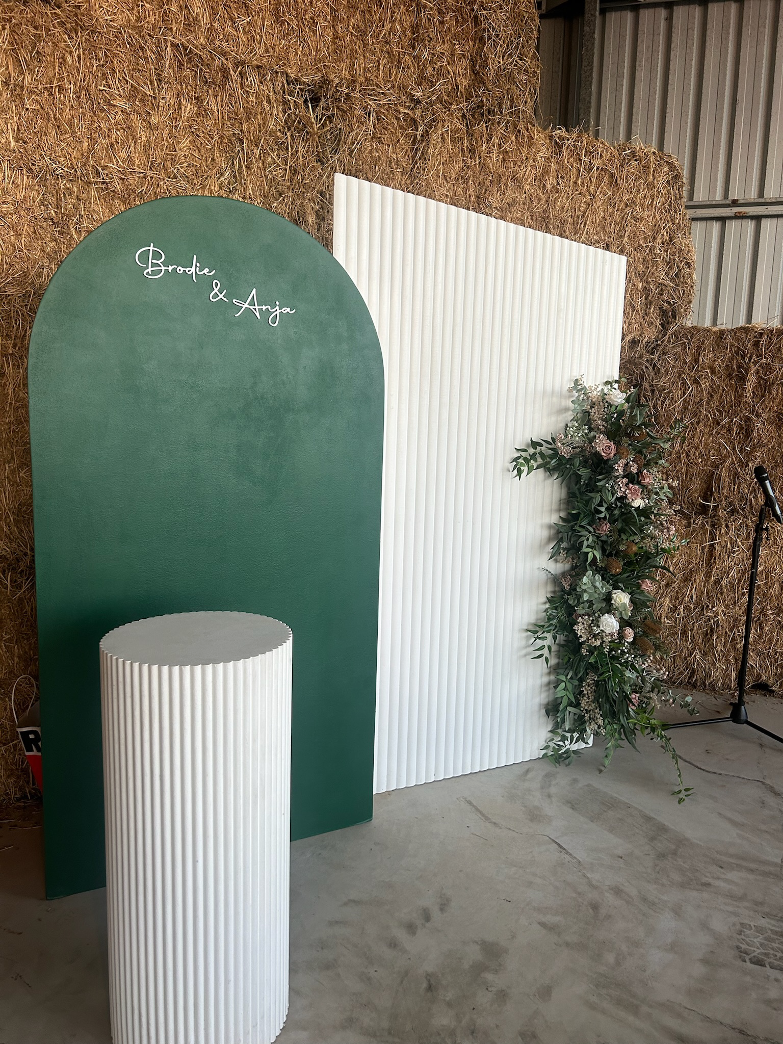 A decorative backdrop with green and white panels, a floral arrangement on the right, and a microphone stand on the far right, set up for a special event.