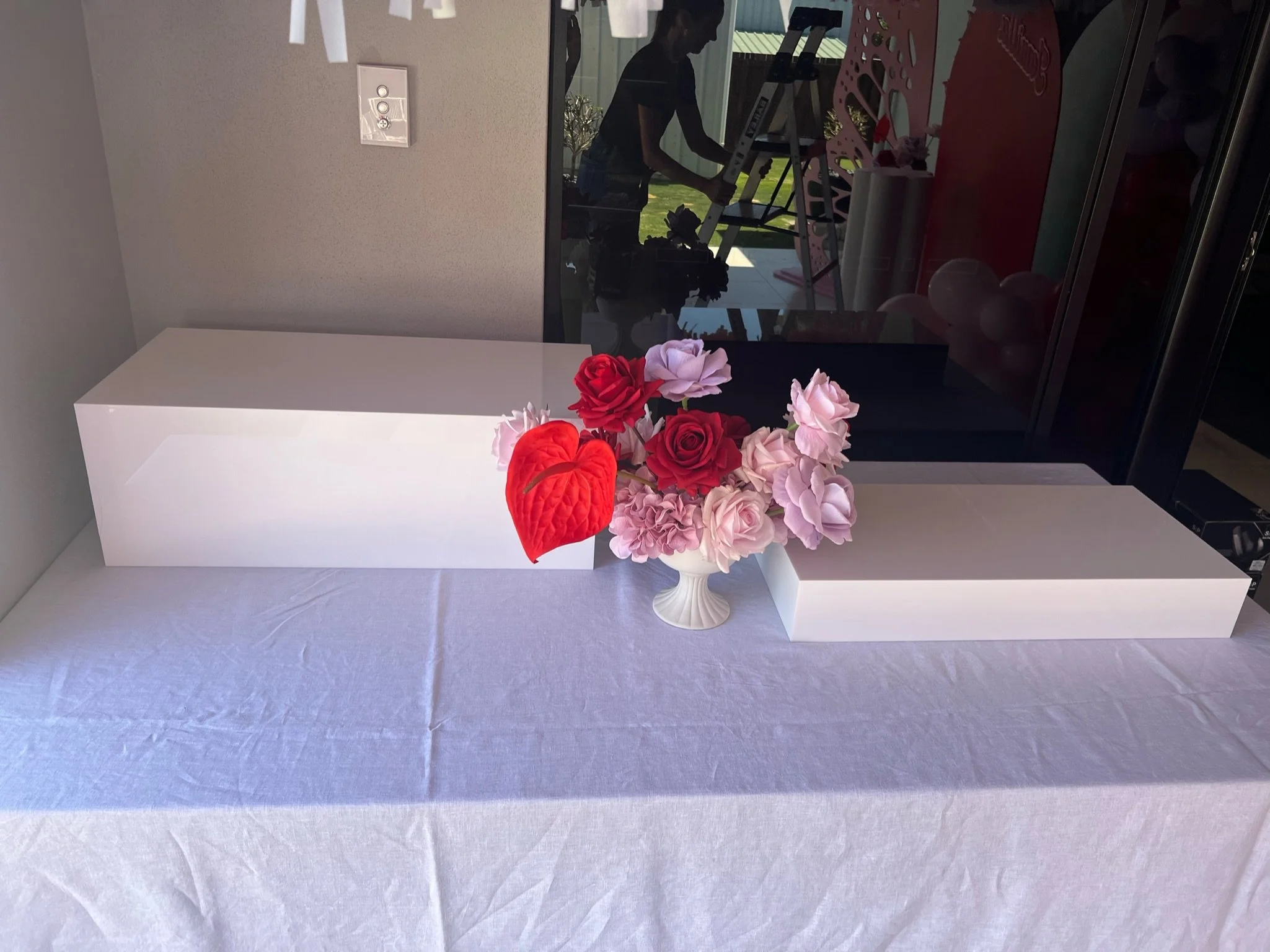 A pink floral arrangement in a white vase with red roses and pink hydrangeas, placed between two white rectangular boxes on a white table with a textured cloth. In the background, a person on a ladder is visible through a glass window.