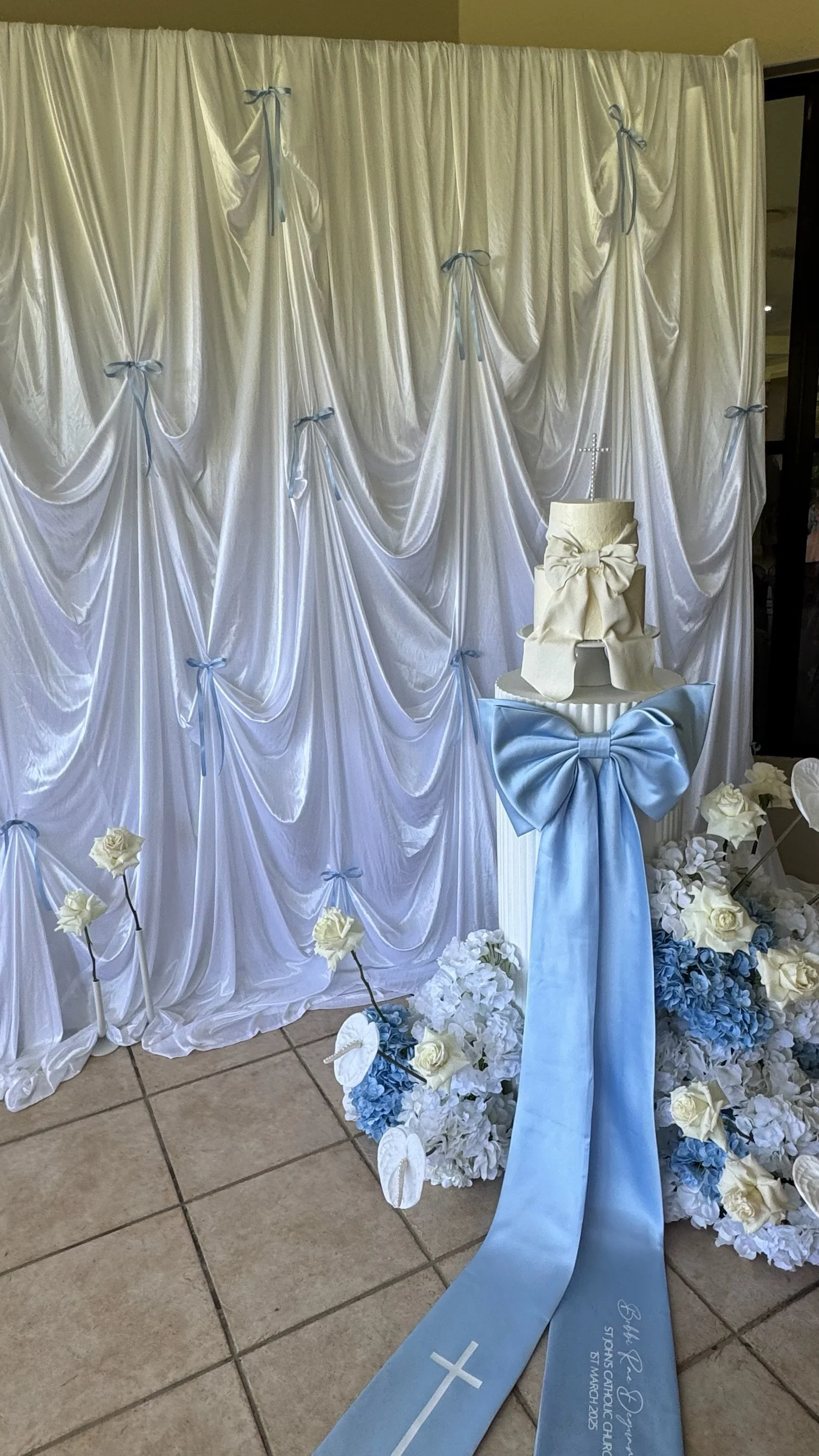 A wedding altar decorated with white curtains tied with blue ribbons, surrounded by white and blue flowers. A celebration cake with a large bow and cross decoration stands on a pedestal draped with a blue satin ribbon.