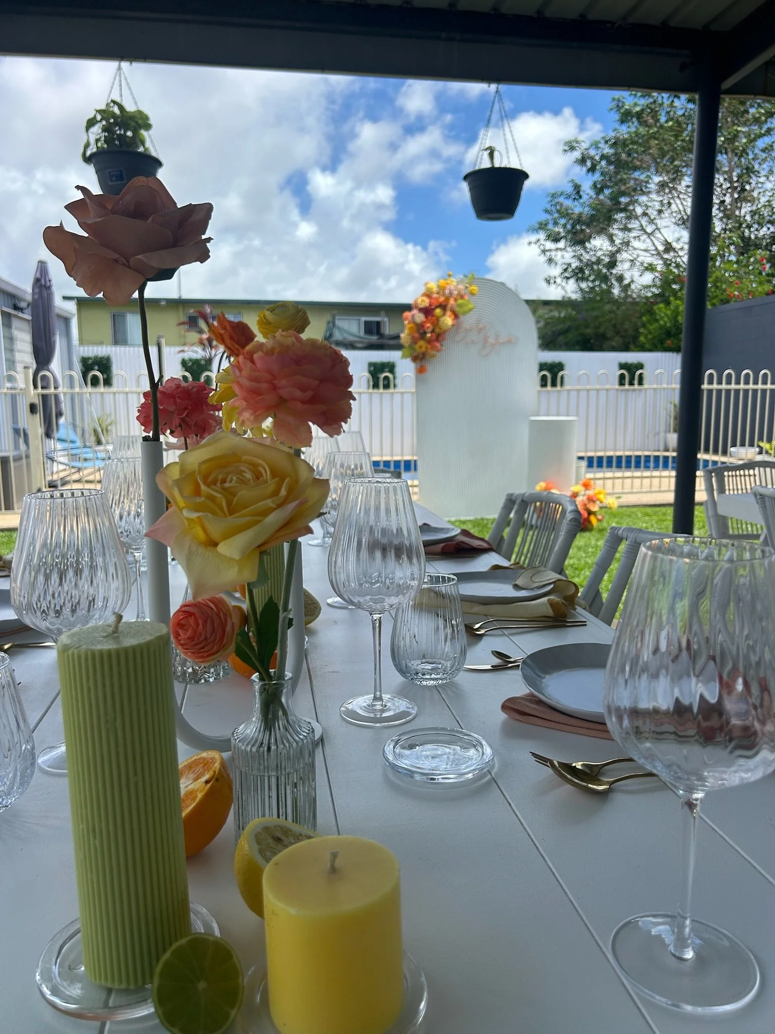 A decorated outdoor table set for a celebration with flowers, candles, and glassware, near a swimming pool with a white fence and a backdrop of a cloudy sky.