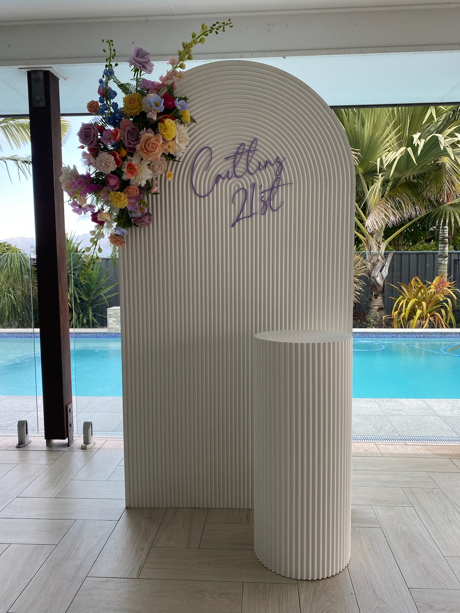 Decorative backdrop with a flower arrangement on the top left, a sign reading 'Cutting 21st' on the center, and a white circular table in front, set indoors with a view of a swimming pool and palm trees outside.