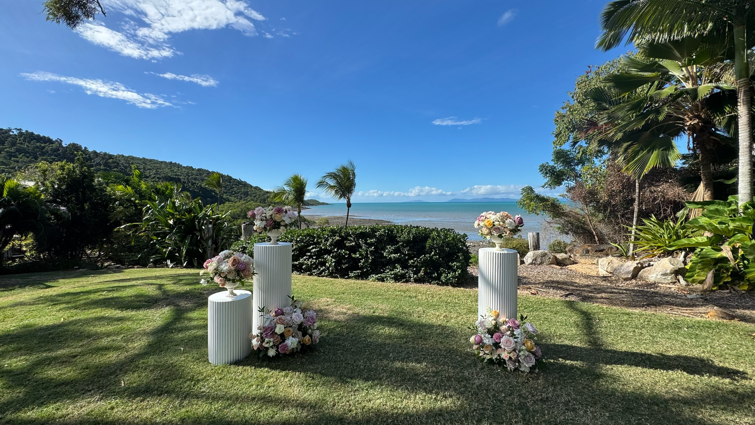Decorative flower arrangements on white pedestals set outside on green grass with ocean, trees, and blue sky in the background.