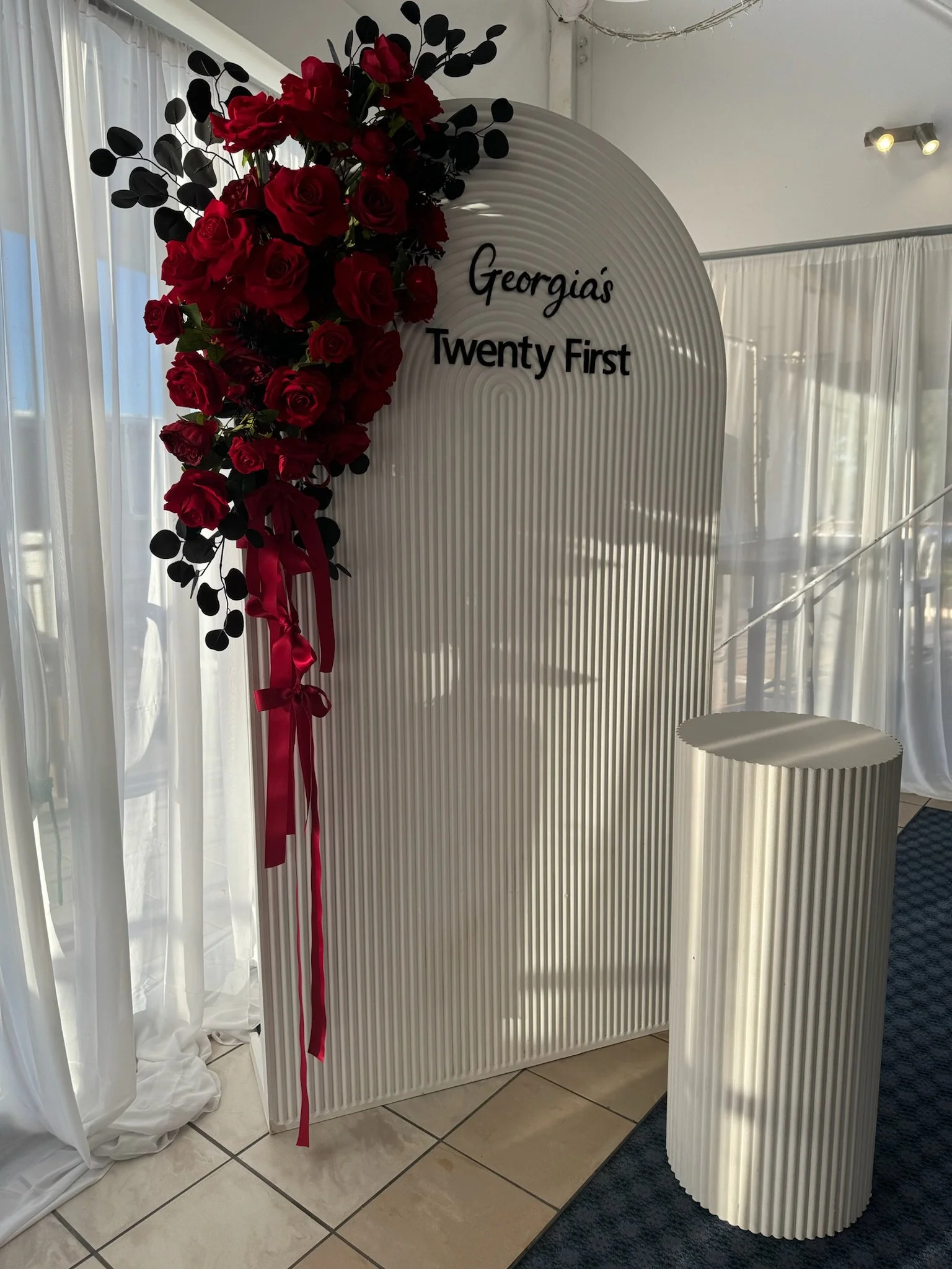 A white, corrugated backdrop with black text that reads 'Georgia's Twenty First'. A floral arrangement of red roses and black leaves is attached to the top left corner, with a red ribbon hanging down. There is a matching white, corrugated cylindrical
