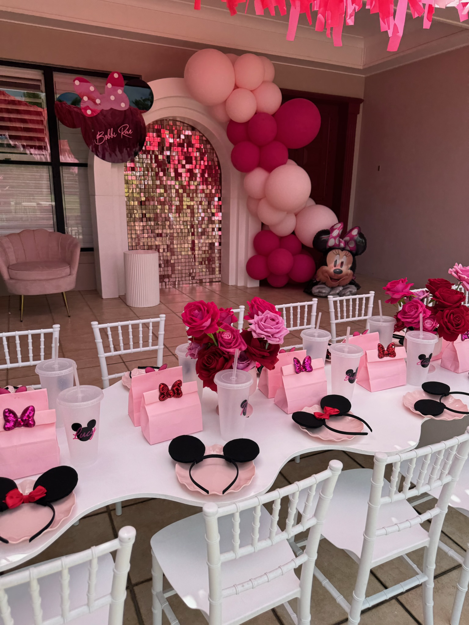 Party decorations with a Minnie Mouse theme, featuring pink and black Mickey ears headbands, pink gift boxes with Minnie Mouse decals, pink and red roses in vases, and Minnie Mouse and Daisy Duck balloons, including a large Minnie Mouse face balloon 