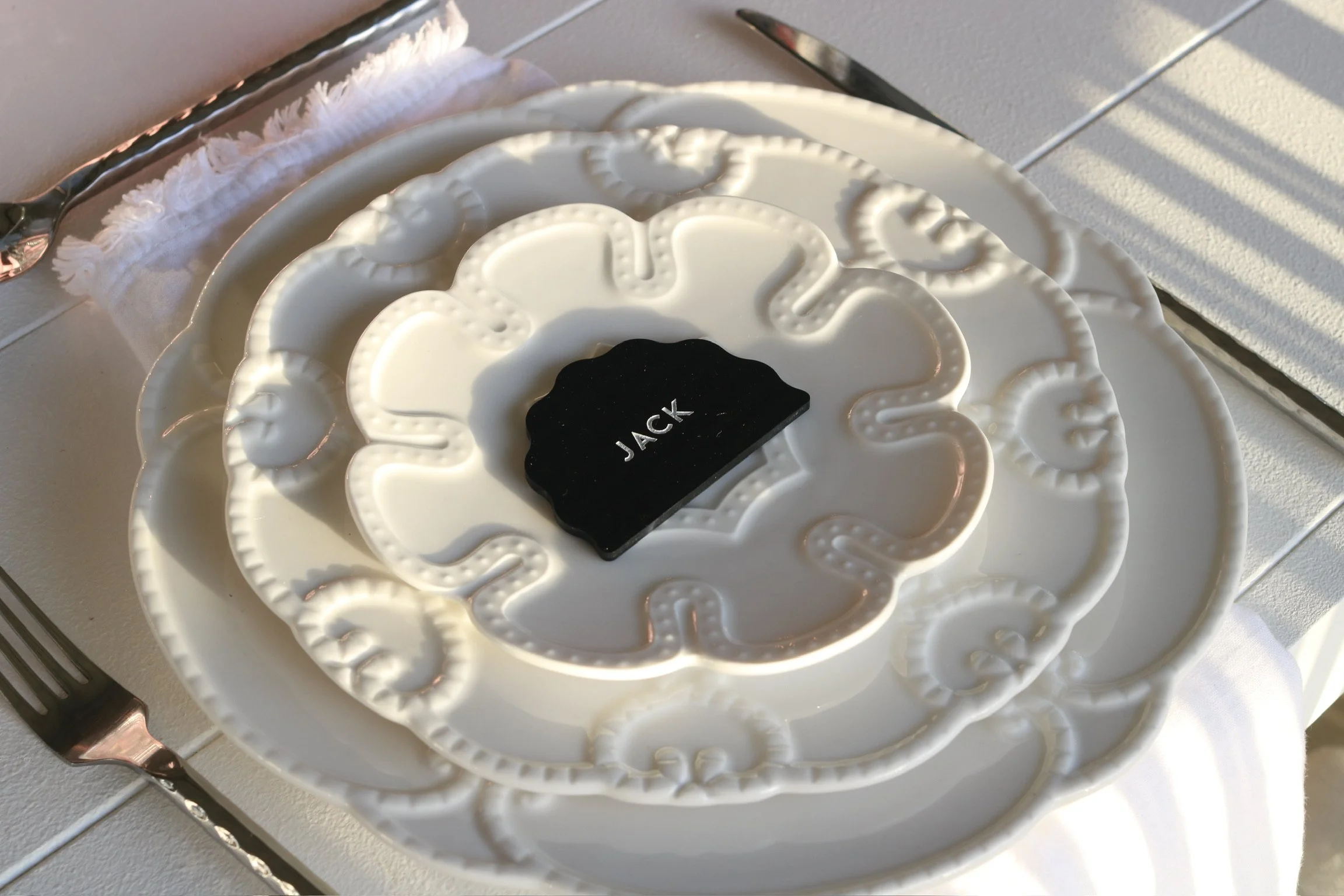 A white decorative plate with a black label that says 'JACK' placed in the center, sitting on a larger white plate with ornate patterns. The setting includes a fork on a white tablecloth, with sunlight casting shadows.