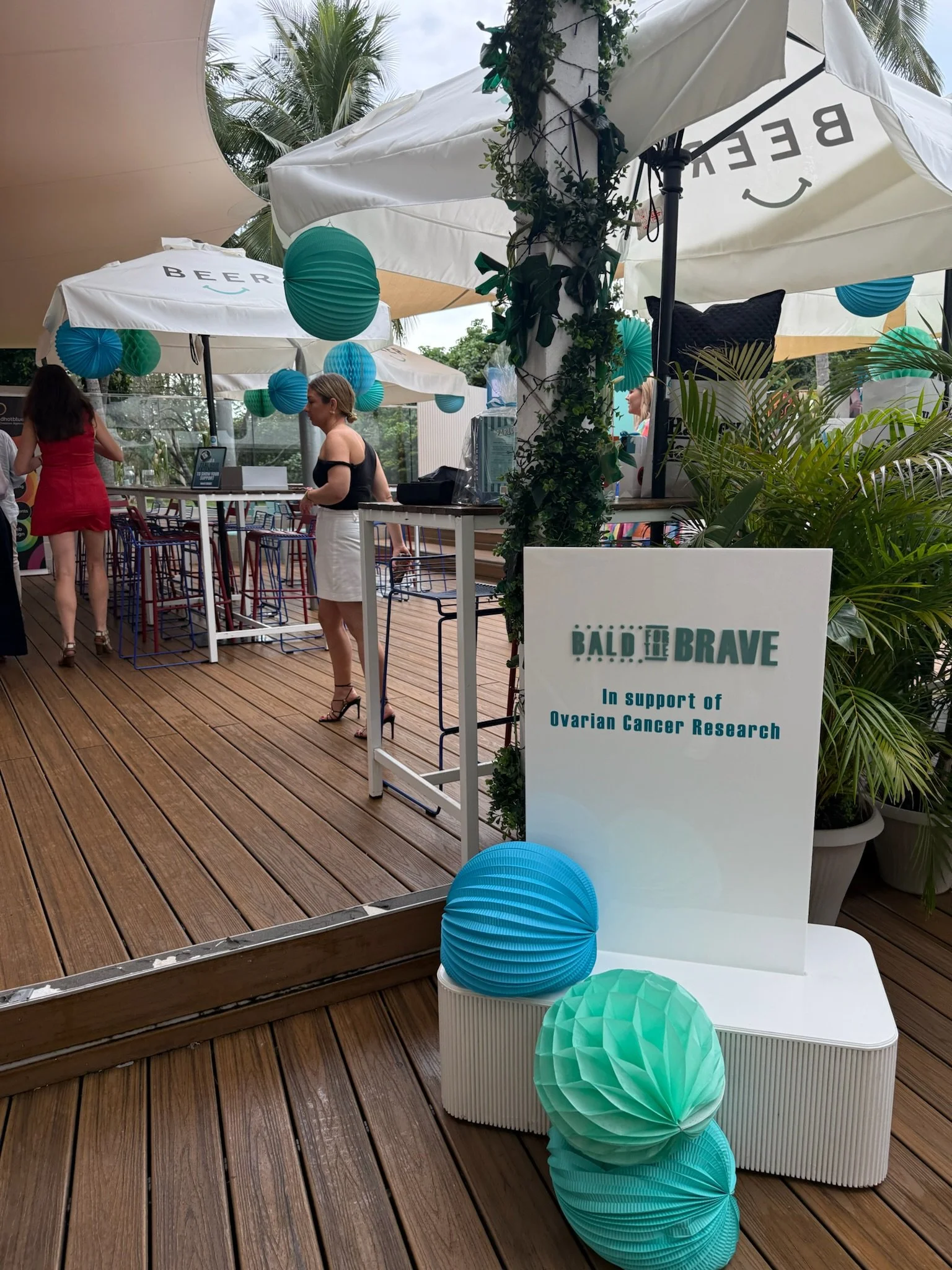 A fundraising event on a rooftop patio with decorated lanterns, a sign supporting ovarian cancer research, and people gathered around tables and bar area.